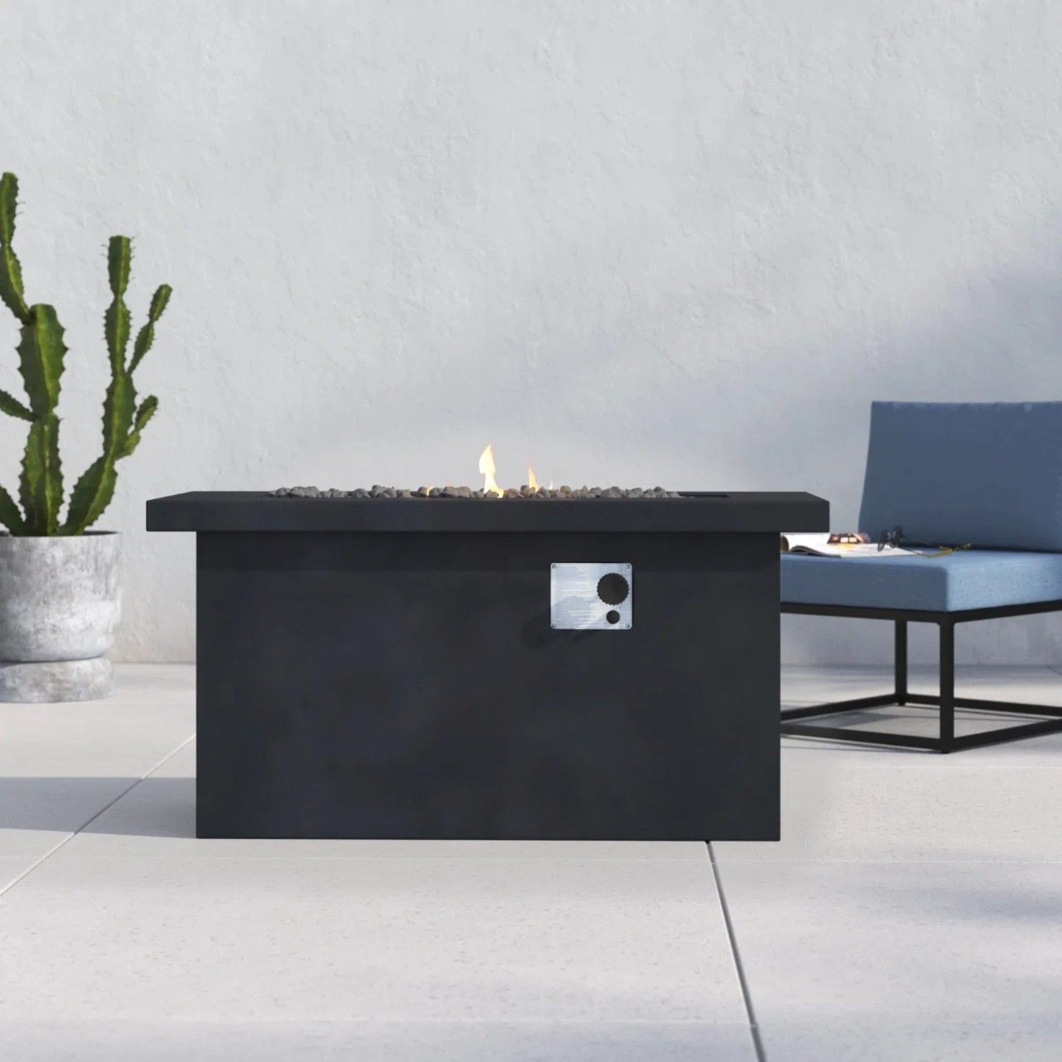 A sleek Propane / Gas Outdoor Fire Pit Table with a small flame sits on a light patio, near a potted cactus and a blue cushioned bench.
