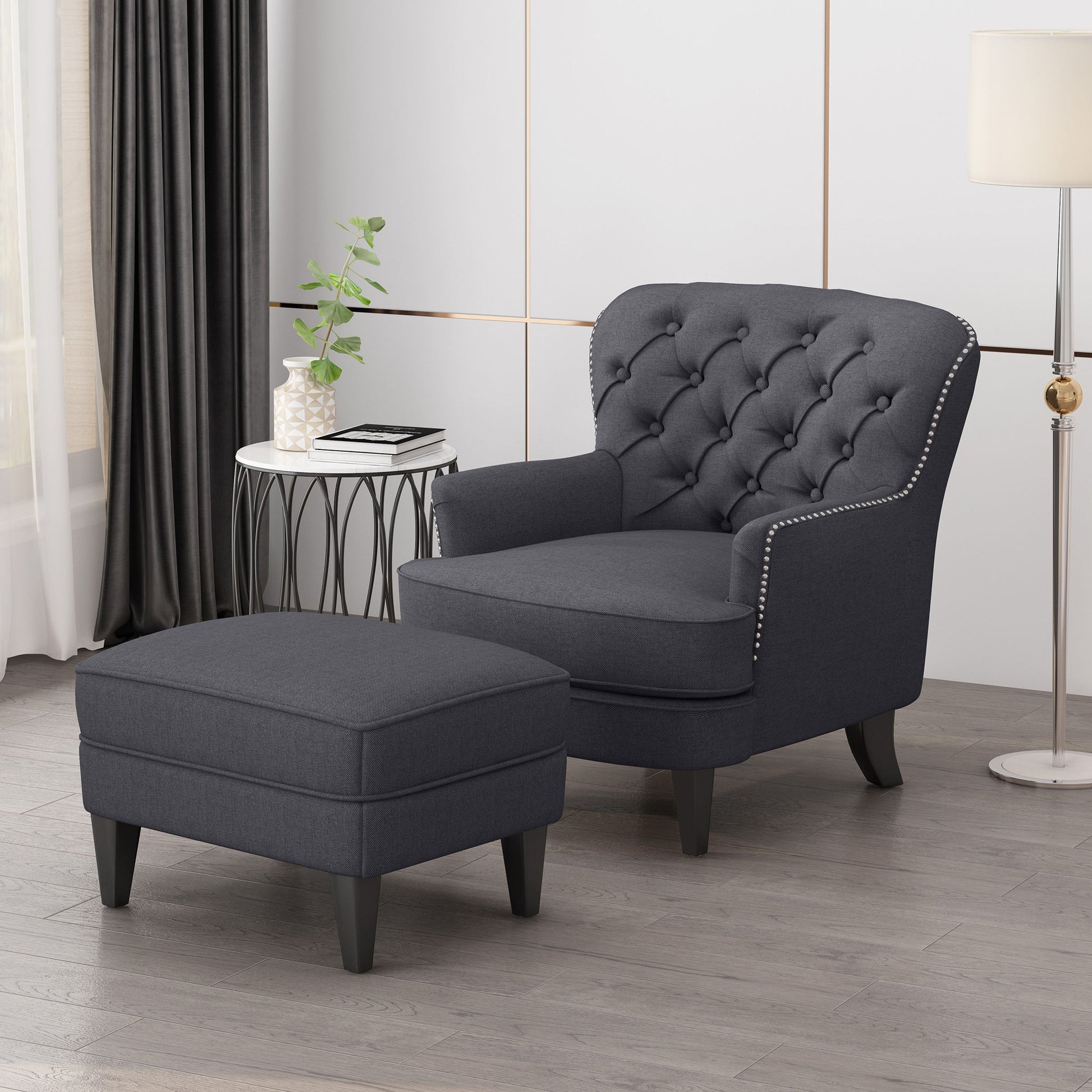 The Enchante Living Room Set, featuring a tufted back club chair and ottoman in dark gray with silver stud trim, is displayed on a wood floor beside a round side table holding a plant, books, and a white lamp in a bright modern living room.