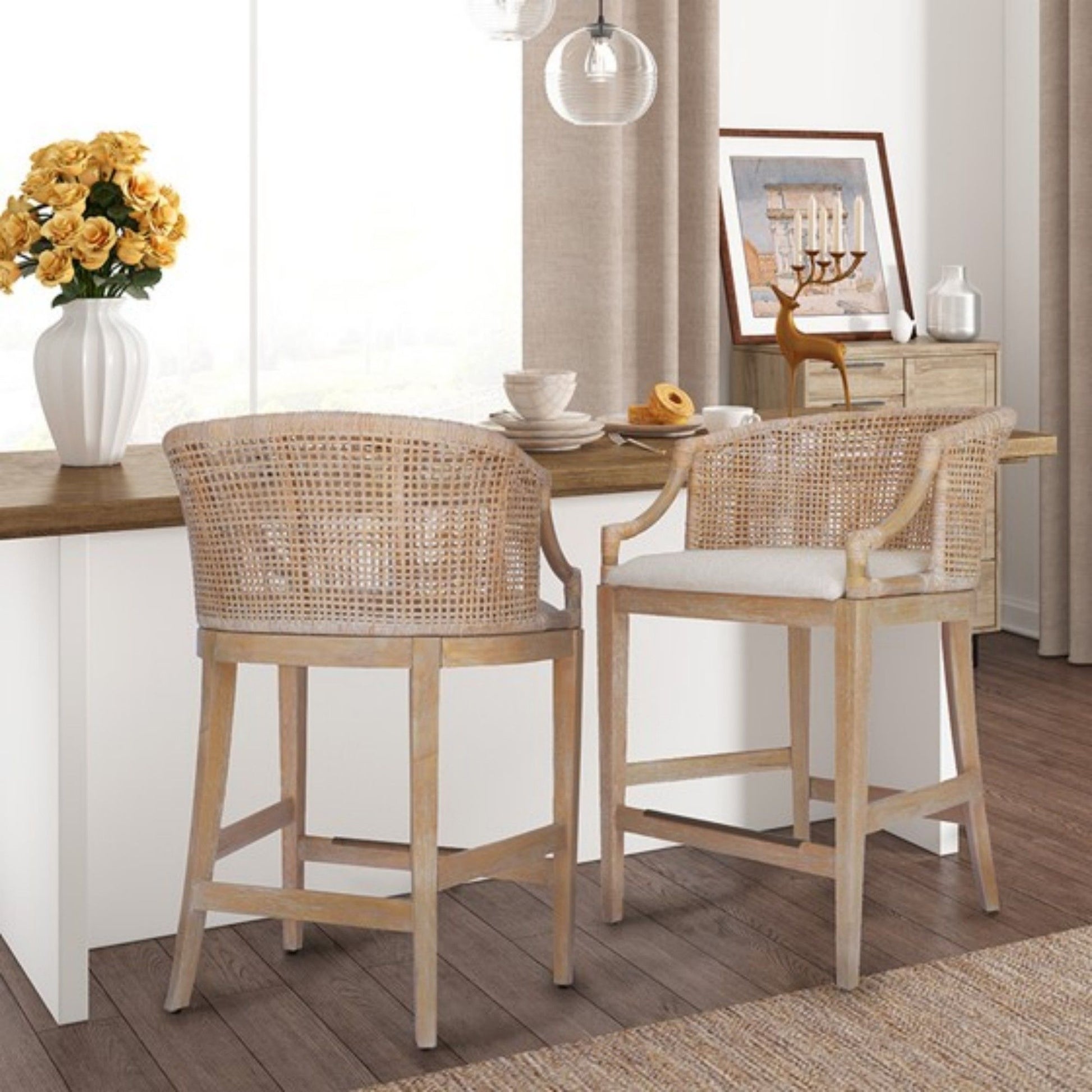 Two Casual Handcrafted Rattan Counter Stools with woven backs and light cushions sit at a breakfast bar in a cozy, sunlit kitchen. Nearby surfaces feature a vase of yellow flowers and decorative accents.