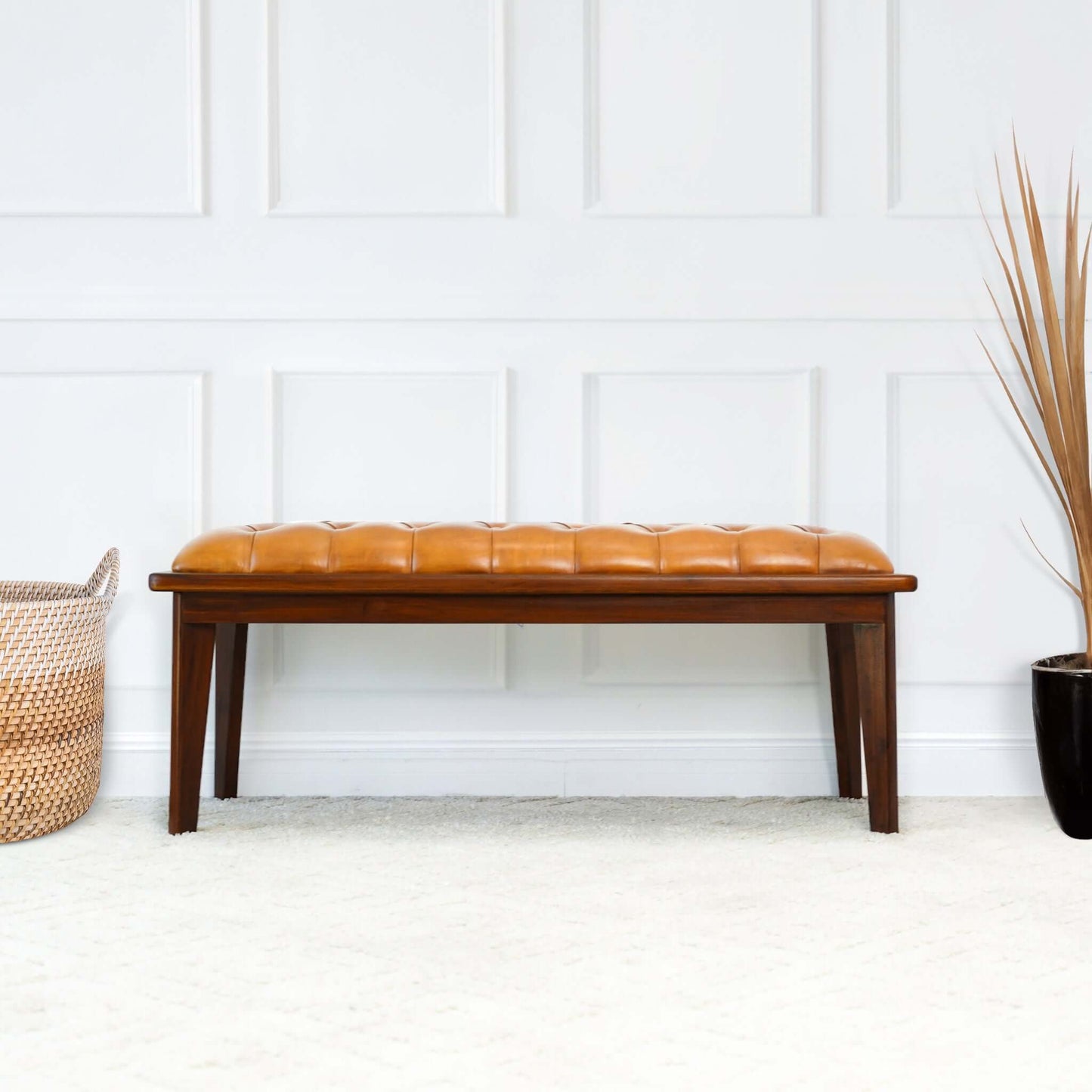 The Arden - Bench With Buttons in brown, rustic tan sits on a light carpet near a white paneled wall, flanked by a woven basket on the left and a black pot with dried grass on the right.