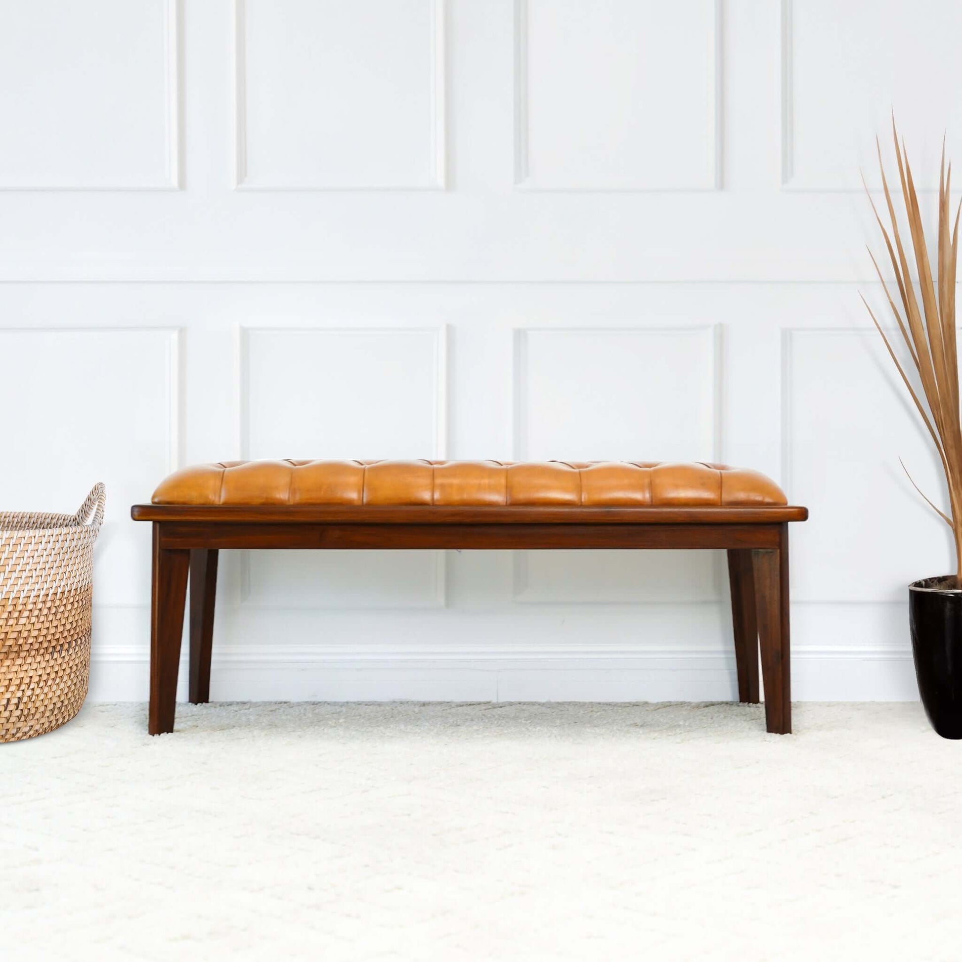The Arden - Bench With Buttons in brown, rustic tan sits on a light carpet near a white paneled wall, flanked by a woven basket on the left and a black pot with dried grass on the right.