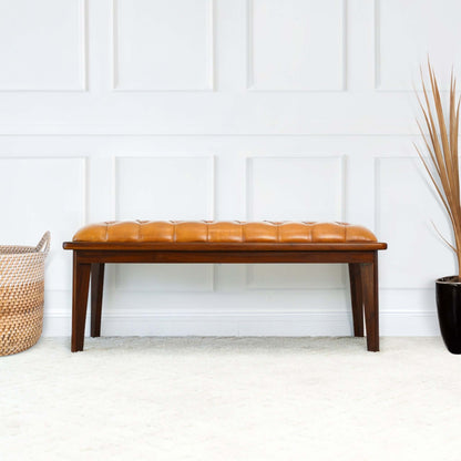 The Arden - Bench With Buttons in brown, rustic tan sits on a light carpet near a white paneled wall, flanked by a woven basket on the left and a black pot with dried grass on the right.