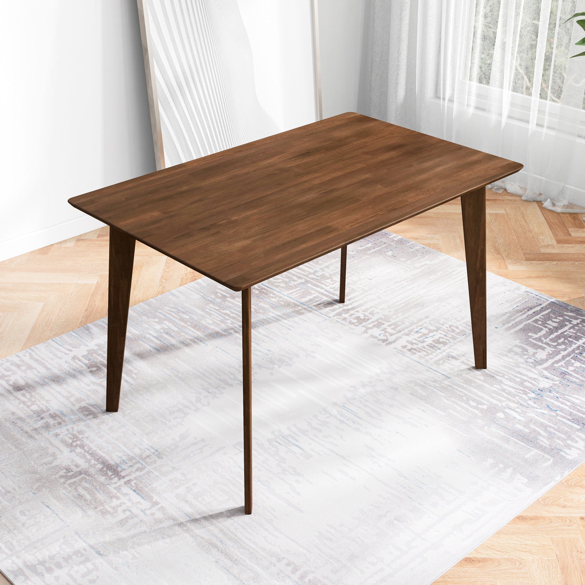 The Carlos Dining Table, featuring a modern wooden design and angled legs, stands on a light-patterned rug in a bright room with large windows, sheer curtains, and a framed artwork against the wall.