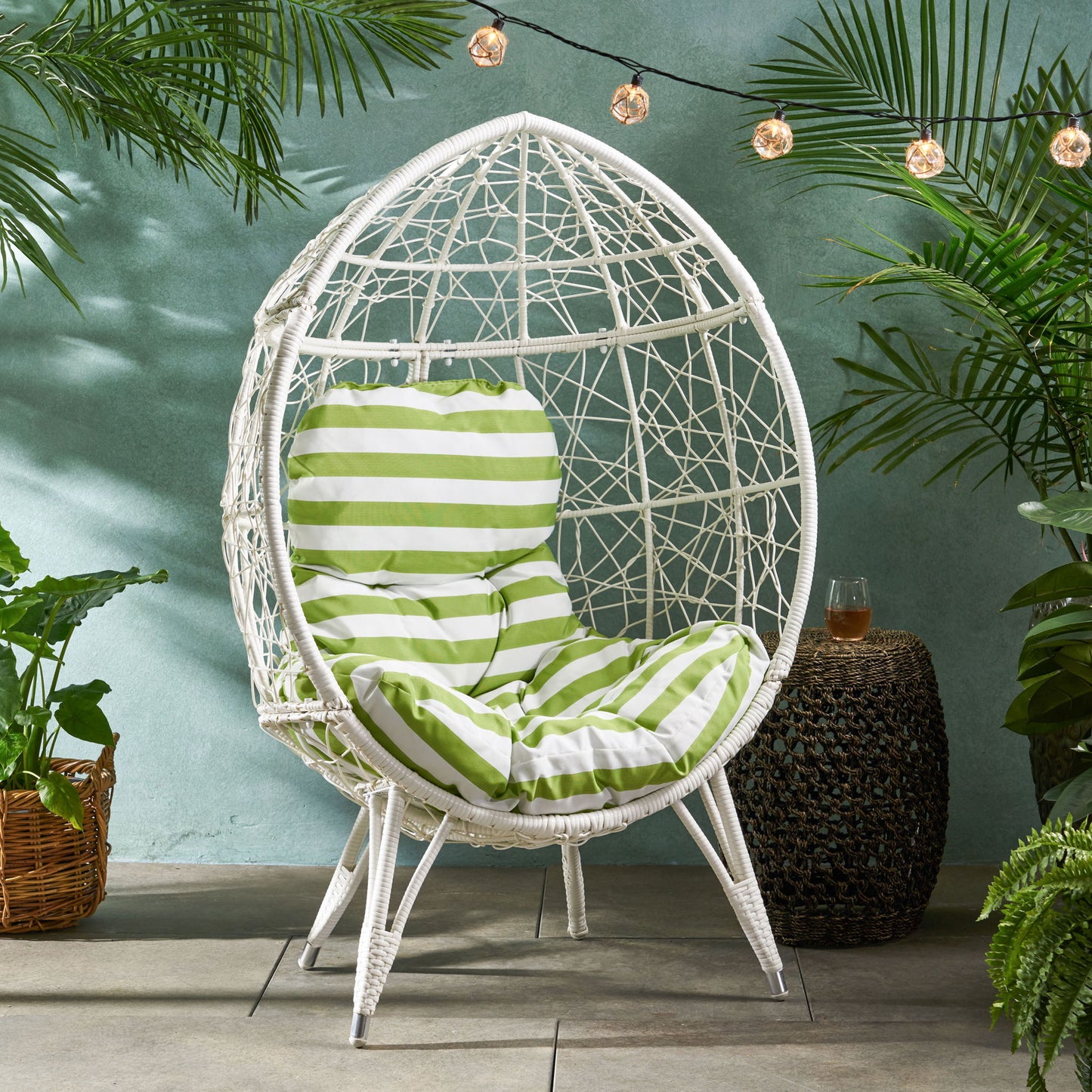 The Cosmos - Round Hammered Iron Teardrop Chair with green and white striped cushions sits on a patio amid tropical plants, beneath string lights. A wicker side table with a drink is nearby.