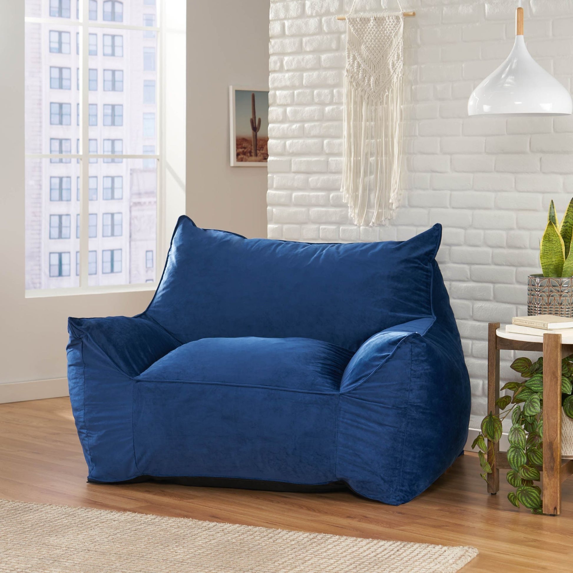 The Allea - Bean Bag Chair With Armrests, in plush blue, is placed in a modern living room featuring white brick walls, a wooden floor, a small round table with plants, and a window overlooking city buildings.