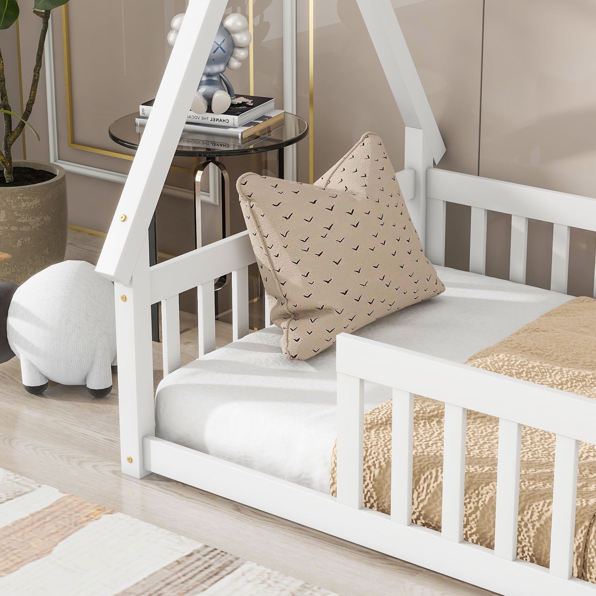 A white Tent-Shaped Floor Bed with guardrails and slats features a beige blanket and patterned pillow. Next to the bed are a round white plush ottoman, potted plant, and side table with books and decor in a cozy room.