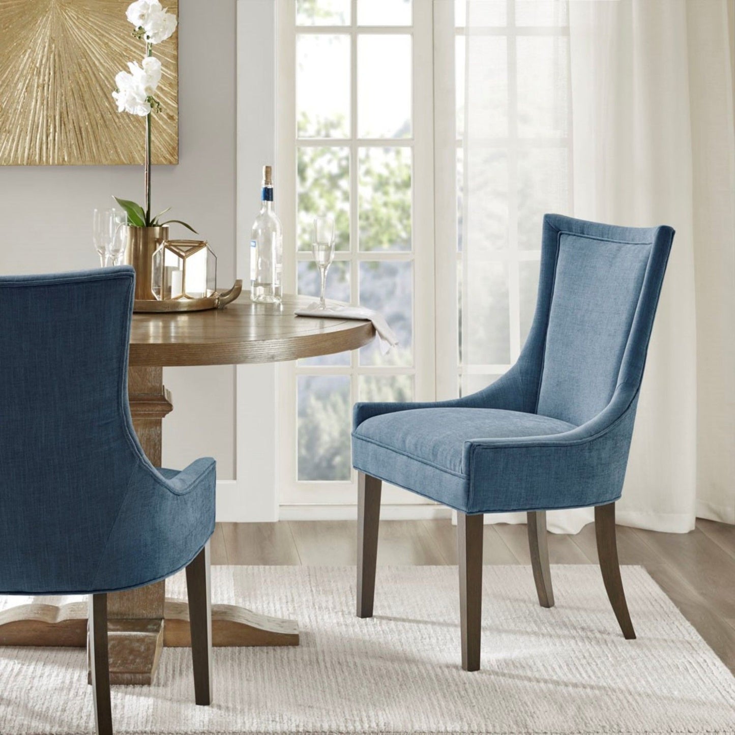 A round wooden dining table set with a wine bottle, glasses, and a vase is surrounded by two Ultra - Elegant Dining Side Chairs on a light rug in a bright room with large windows and sheer curtains.
