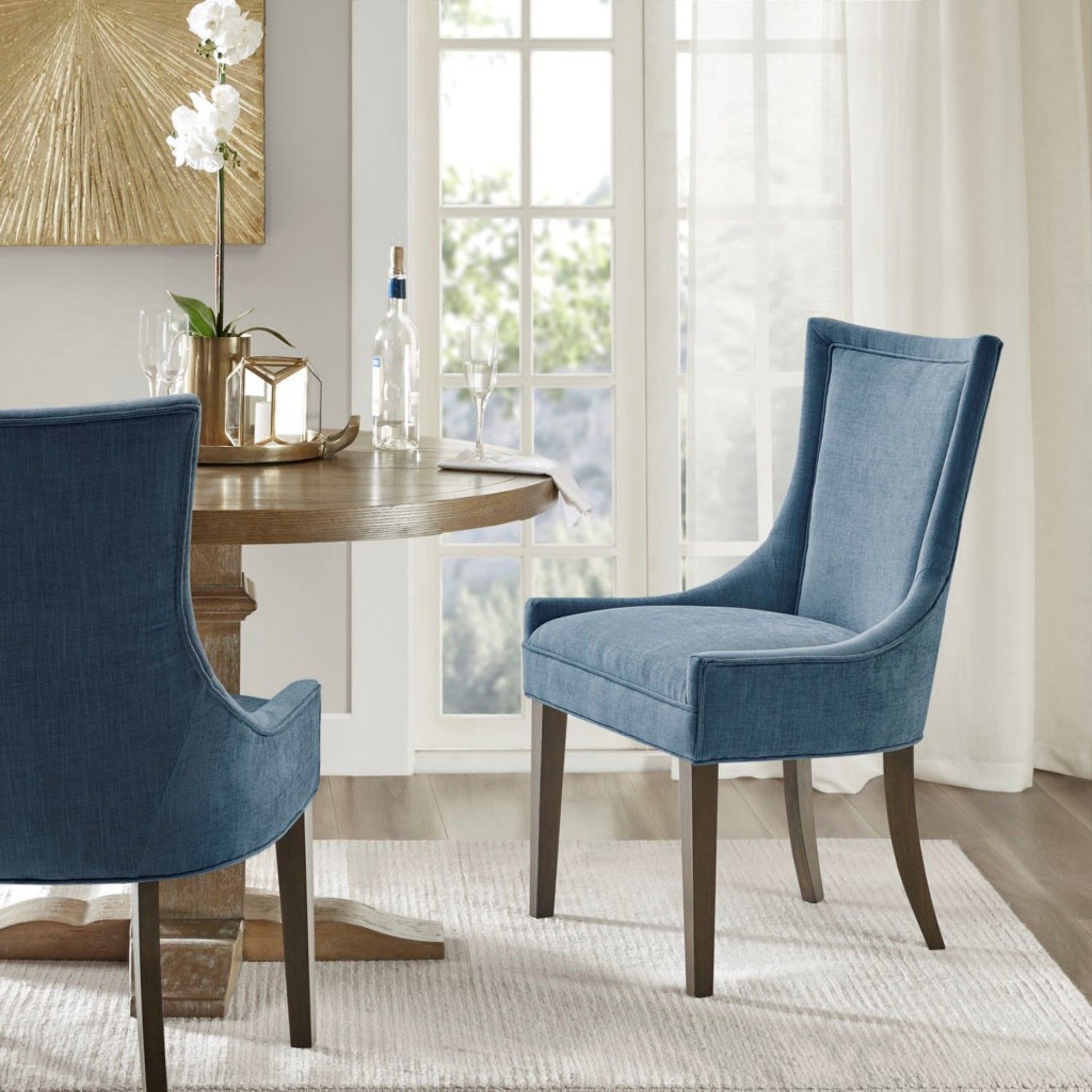 A round wooden dining table set with a wine bottle, glasses, and a vase is surrounded by two Ultra - Elegant Dining Side Chairs on a light rug in a bright room with large windows and sheer curtains.