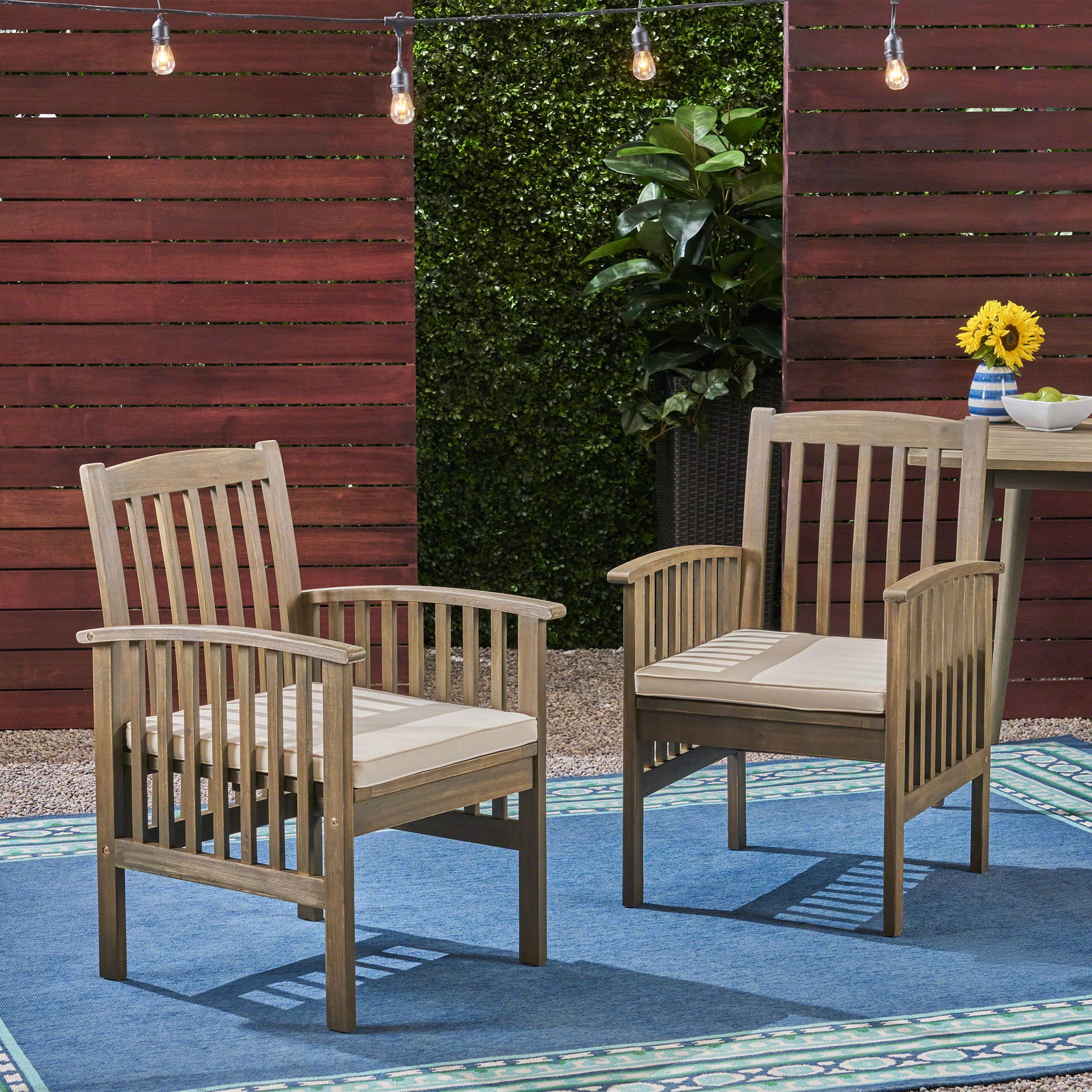 Two Casa Acacia Dining Chairs with beige cushions sit on a blue outdoor rug, under string lights. A table with sunflowers and a potted plant are in the background, framed by wooden fencing and green hedges.