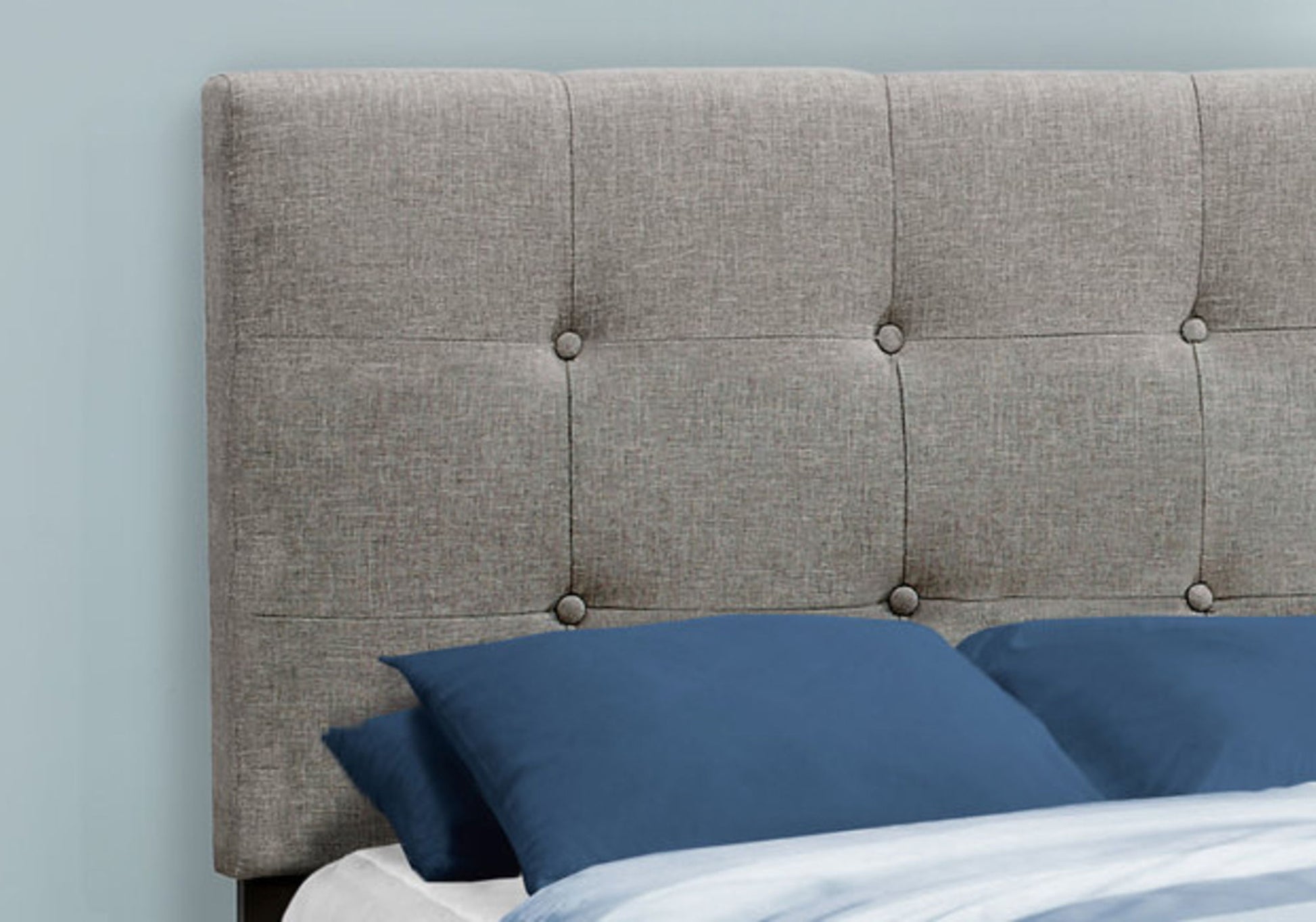 A close-up of the Upholstered Bed Frame Only, featuring a gray tufted headboard, behind blue pillows and a light blue comforter, set against a pale blue wall.