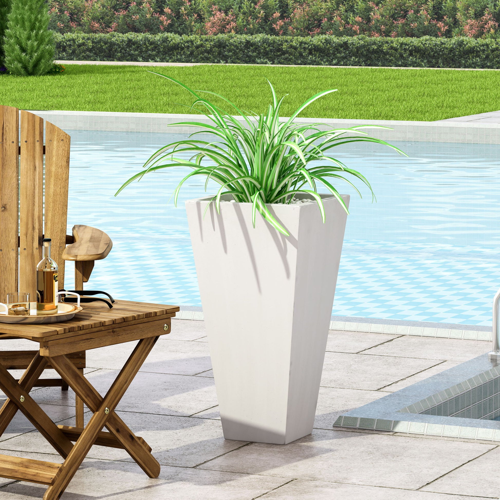 An Outdoor Modern Cast Stone Planter with long, green leaves sits on a poolside patio next to a wooden chair and table, with the pool and green lawn in the background.