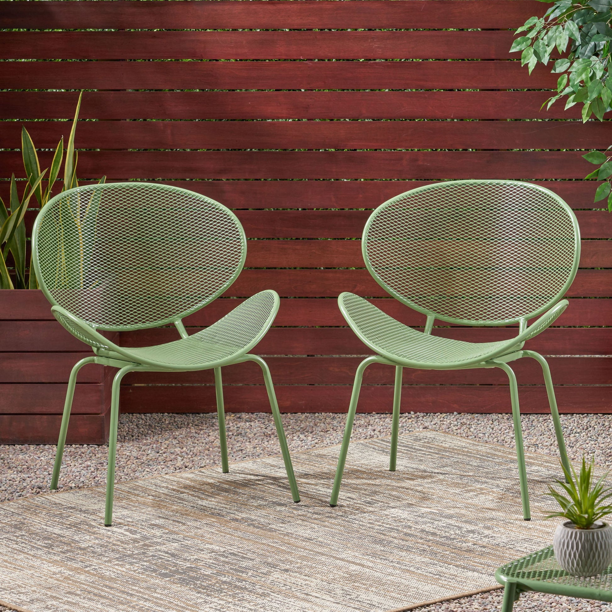 Two Elba Contemporary Chairs, featuring modern green metal frames and mesh backs and seats, sit on a beige outdoor rug surrounded by gravel, potted plants, and a wooden privacy fence.