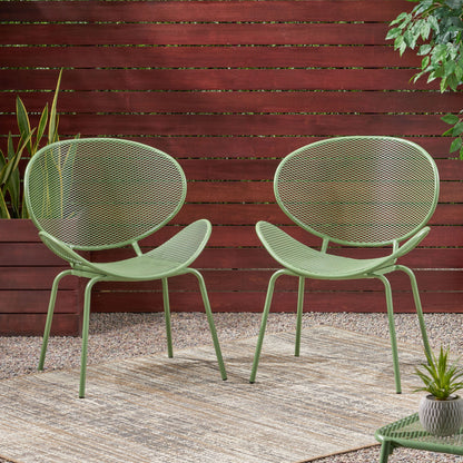 Two Elba Contemporary Chairs, featuring modern green metal frames and mesh backs and seats, sit on a beige outdoor rug surrounded by gravel, potted plants, and a wooden privacy fence.