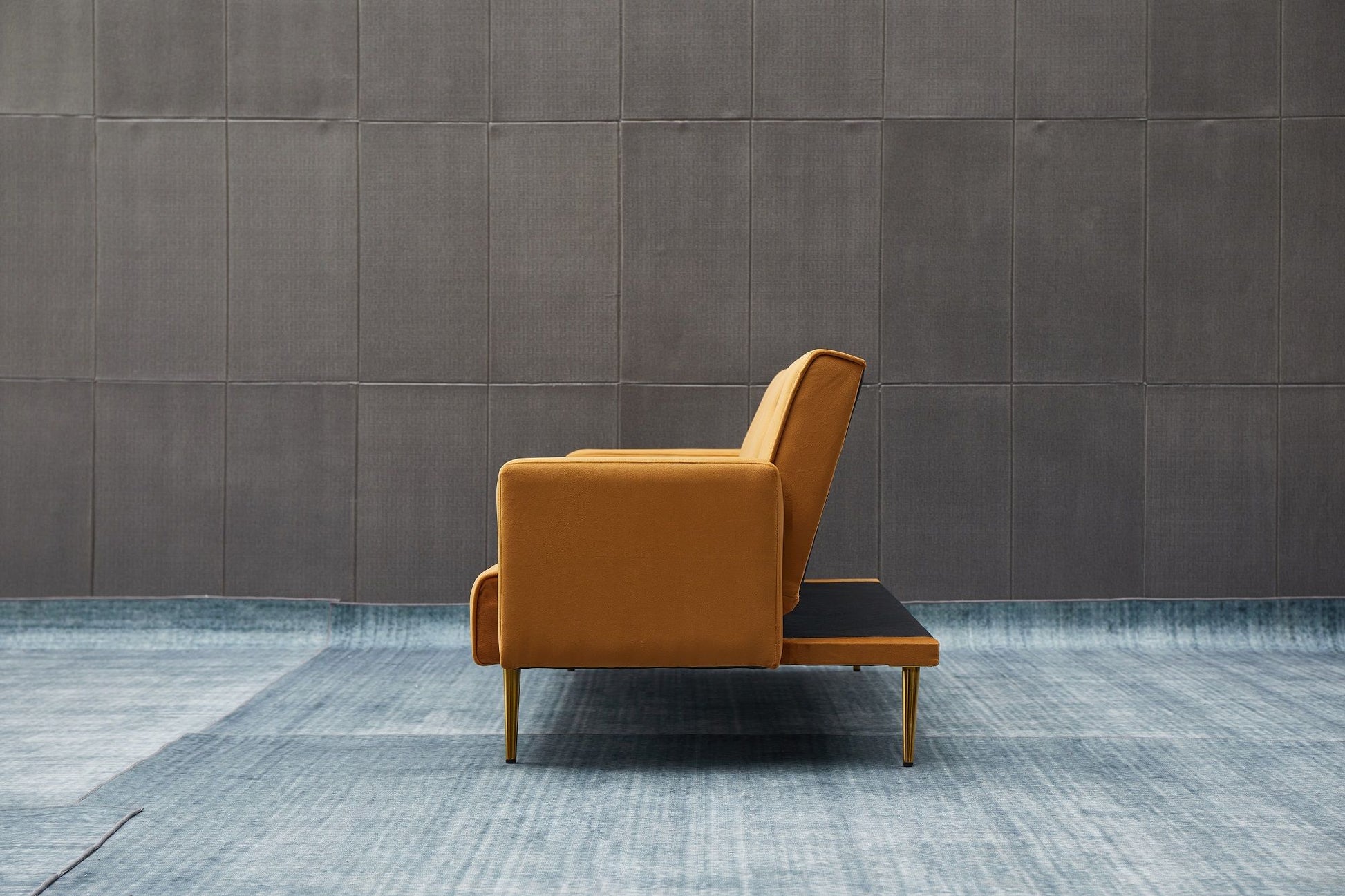 The Comfort Compact Design Sofa Bed, in modern mustard-yellow with wooden legs, is shown in side profile against a gray paneled wall and set on a light blue textured rug.