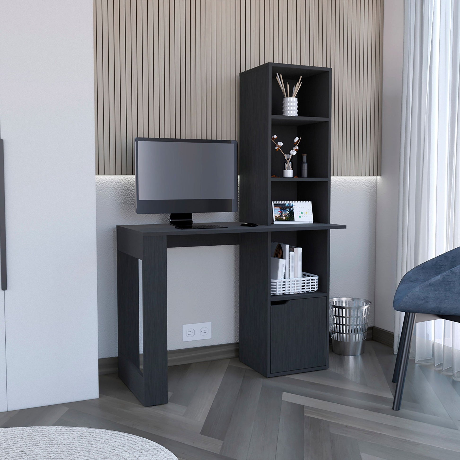 The Writing Desk With Bookcase And Cabinet - Black, placed against a paneled wall by a window with sheer curtains, holds a monitor, books, and decor. A blue chair and metal trash bin are nearby.