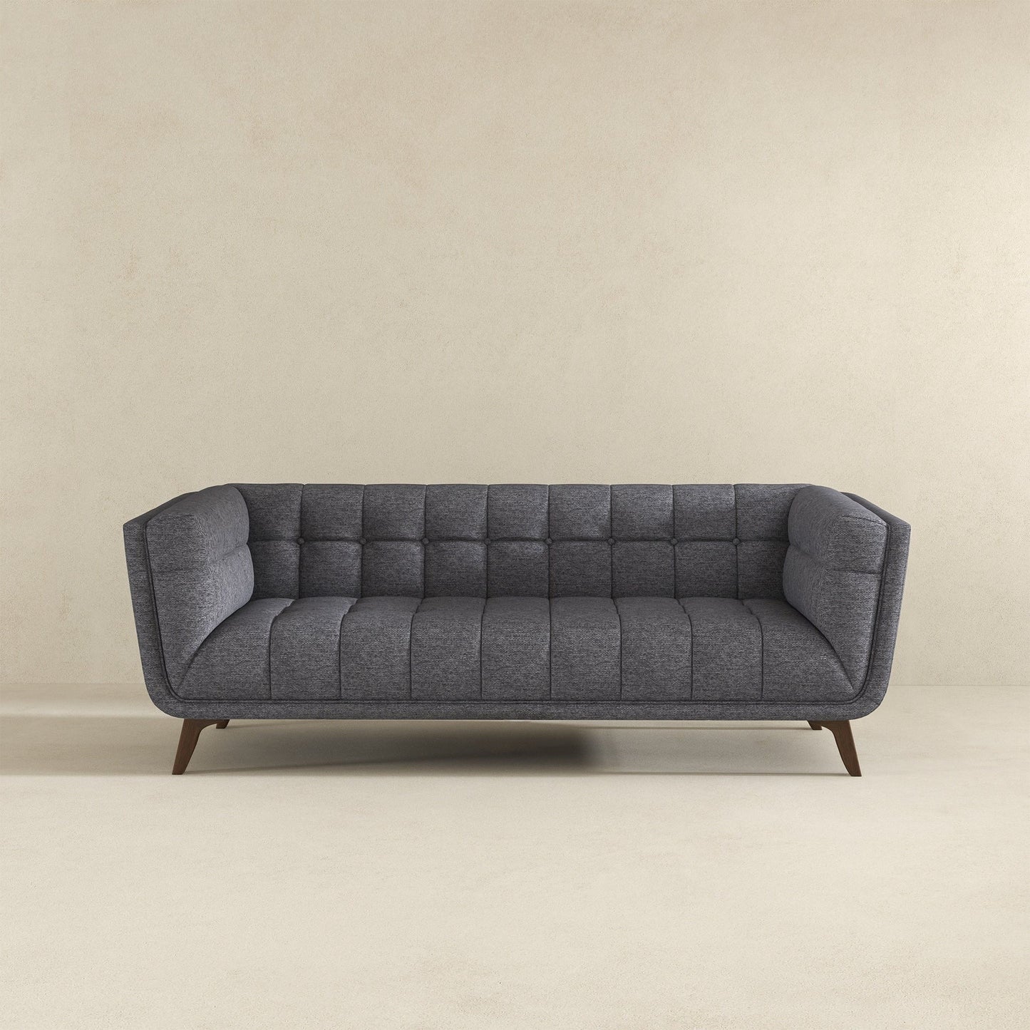 The Addison Mid-Century Modern Tufted Sofa features dark gray upholstery, tufted cushions, and angled wooden legs, shown against a plain beige wall and a light-colored floor.