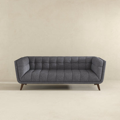 The Addison Mid-Century Modern Tufted Sofa features dark gray upholstery, tufted cushions, and angled wooden legs, shown against a plain beige wall and a light-colored floor.
