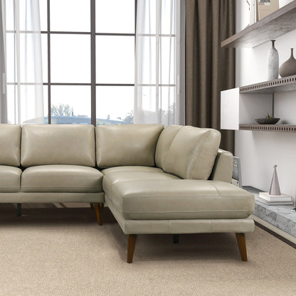 The Benson - Sectional Sofa With Chaise, in light gray leather with wooden legs, sits in a modern living room by a large window with sheer curtains, neutral walls, and minimalist shelves displaying decorative vases.