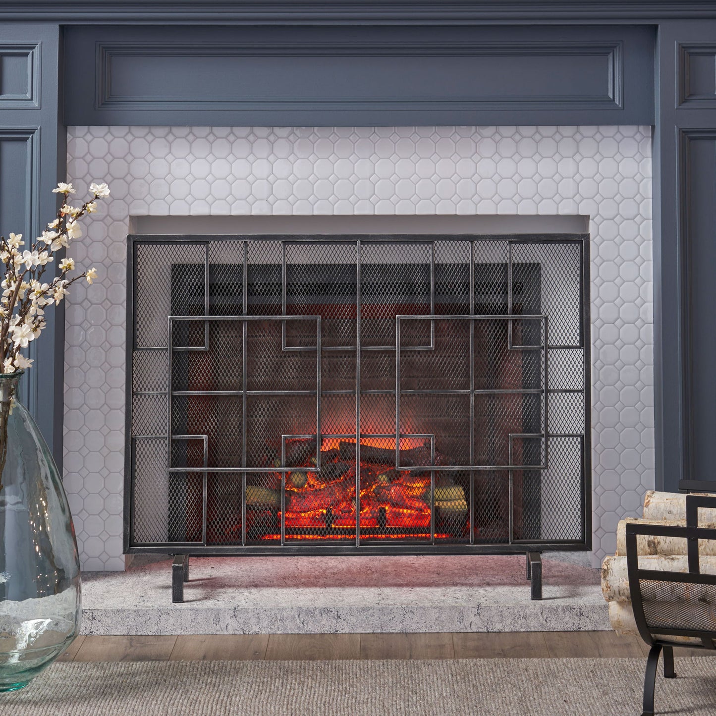A modern living room features the Dorothy - Modern Single Panel Iron Fire Screen, glowing artificial logs, hexagonal white tile surround, a gray mantle, and a large glass vase with white flowers on the left.