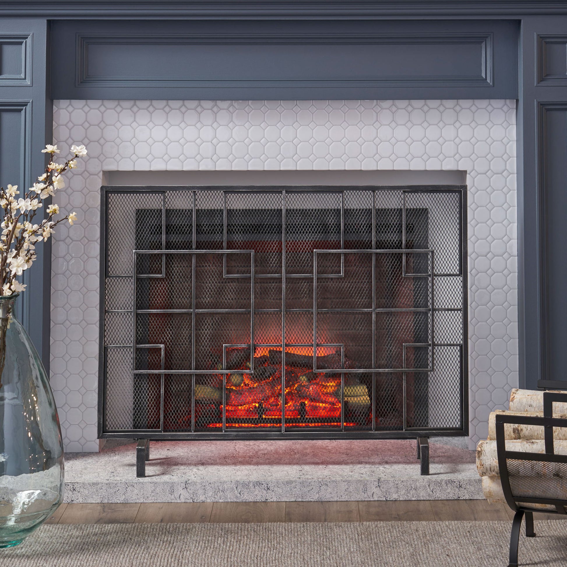A modern living room features the Dorothy - Modern Single Panel Iron Fire Screen, glowing artificial logs, hexagonal white tile surround, a gray mantle, and a large glass vase with white flowers on the left.
