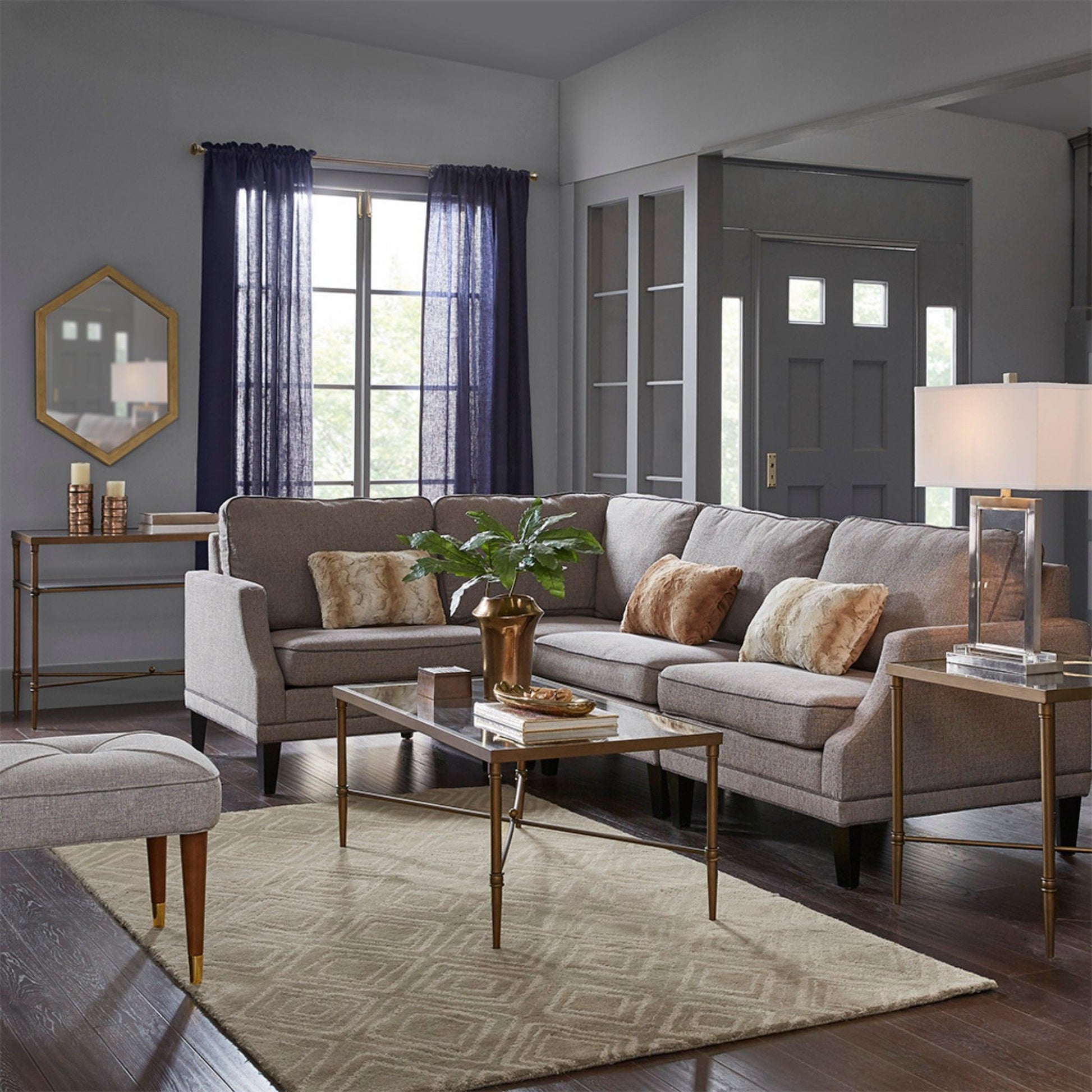 A modern living room with gray sofas, brown and beige pillows, a Metal Rectangle Coffee Table With Tempered Glass Top - Bronze featuring a potted plant, beige rug, dark wood floors, and windows with dark curtains letting in natural light.