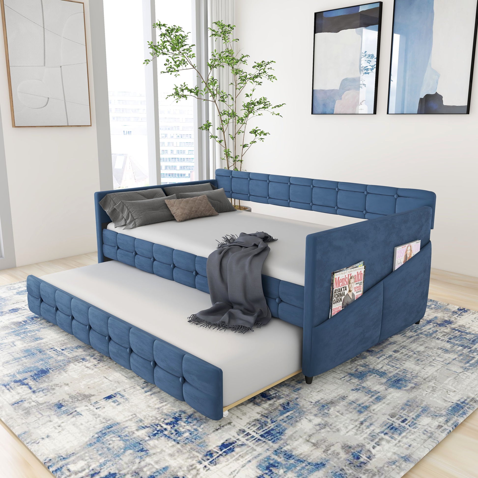 A modern bedroom features the Upholstered Twin Size Daybed Bed Frame with Trundle in blue velvet and studding design. The daybed has a gray blanket, pillows, side pockets for magazines, with abstract art above and a blue-patterned rug below.