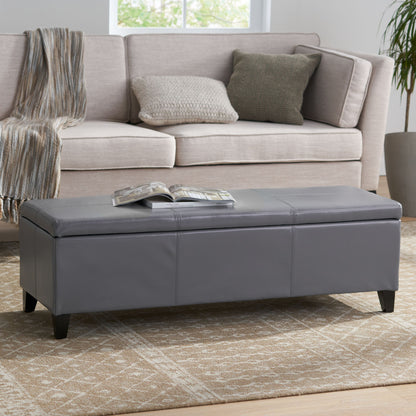 The Glouster - Contemporary Storage Ottoman, upholstered in gray, sits on a patterned rug with a book on top, placed in front of a beige sofa with throw pillows and a striped blanket in a bright living room.