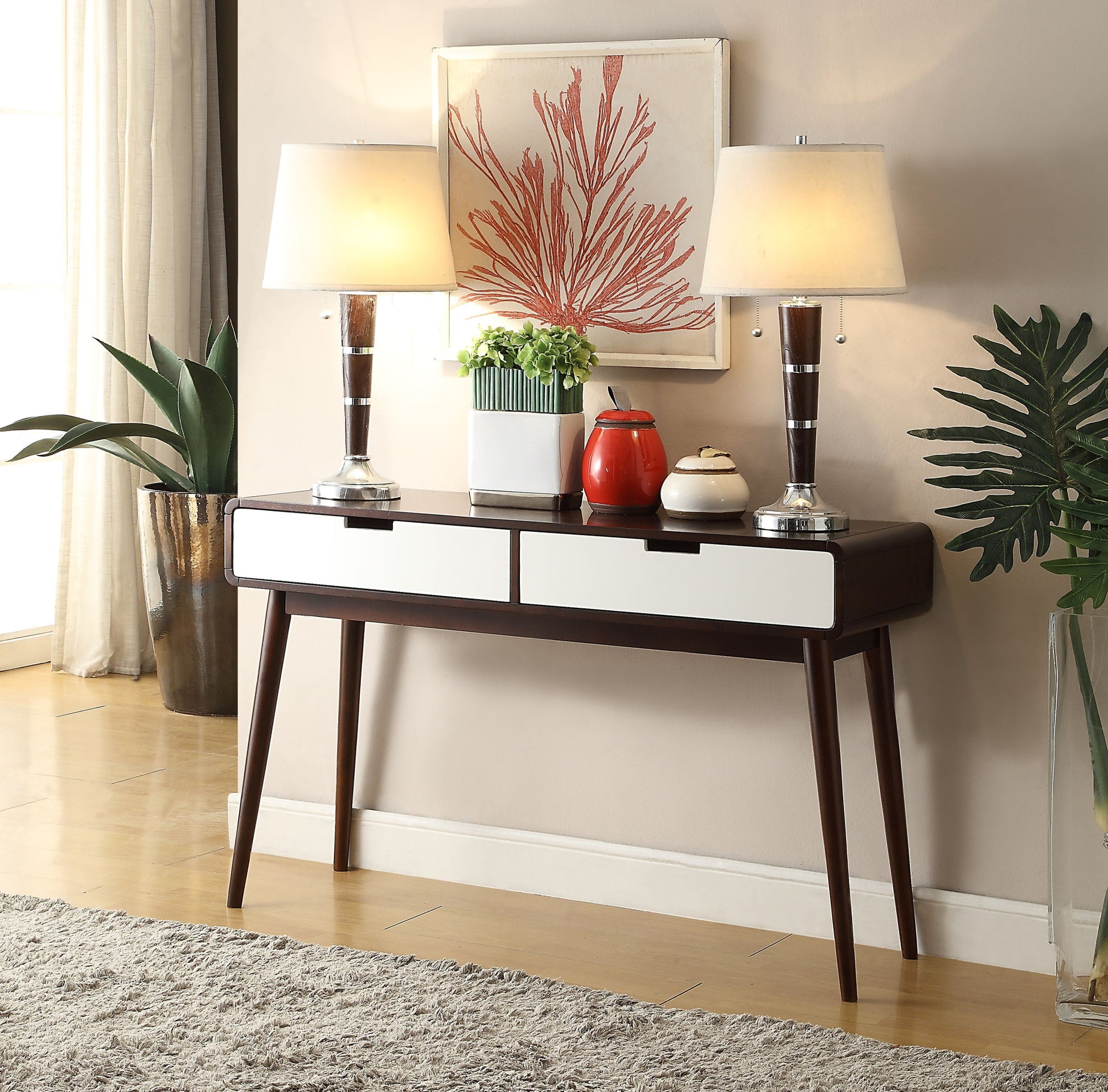 The Christa Sofa Table in White/Espresso features two drawers and displays matching lamps, a red teapot, a plant, and a decorative bowl. Above it hangs coral artwork, flanked by green plants on both sides with a beige rug below.