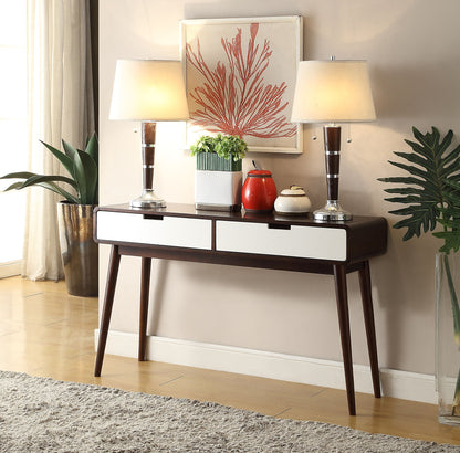The Christa Sofa Table in White/Espresso features two drawers and displays matching lamps, a red teapot, a plant, and a decorative bowl. Above it hangs coral artwork, flanked by green plants on both sides with a beige rug below.
