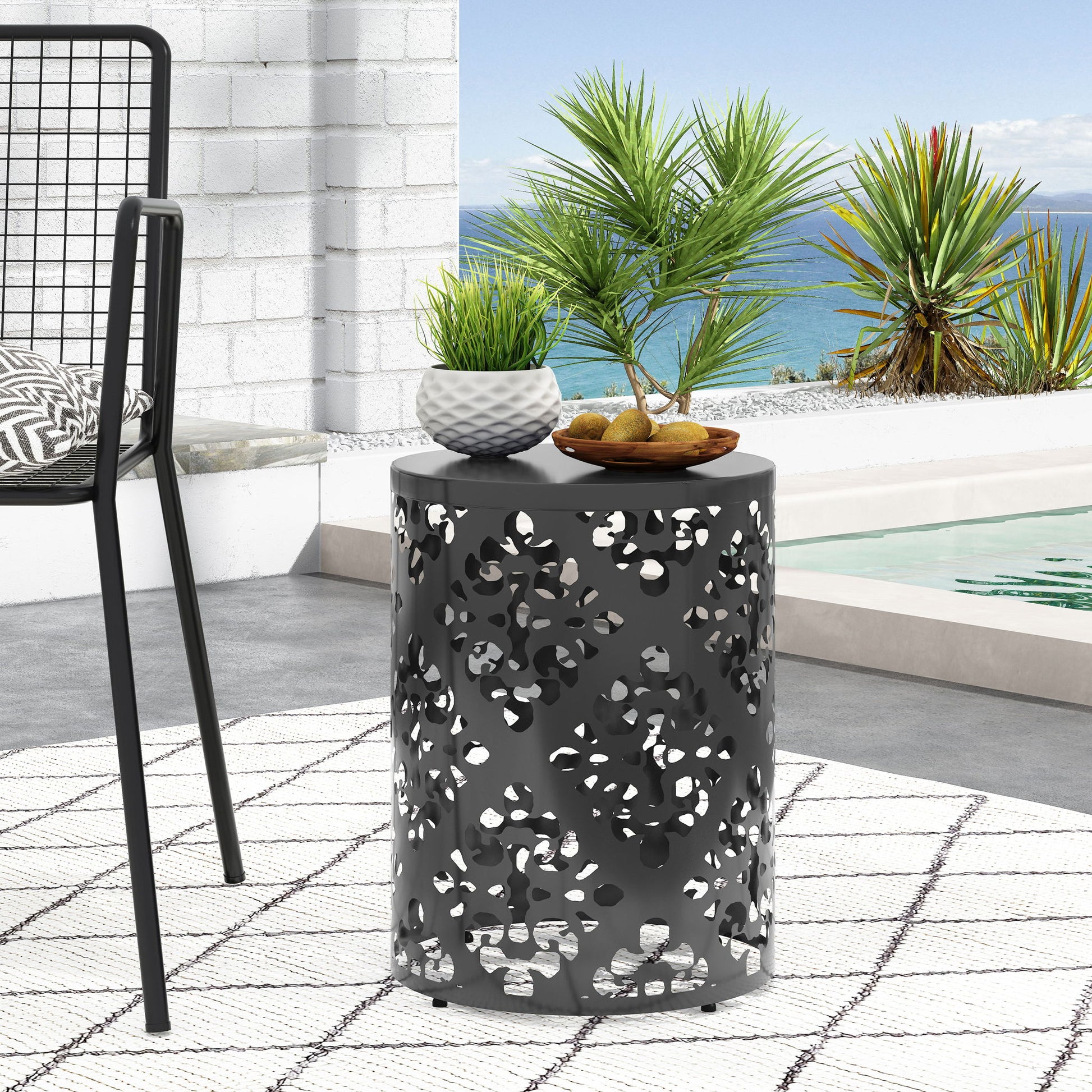 The Outdoor Metal Modern Design End Table, featuring a black finish and floral cutouts, sits on a white patterned outdoor rug beside a pool. Potted plants and fruit decorate the table, with ocean views and tropical plants in the background.
