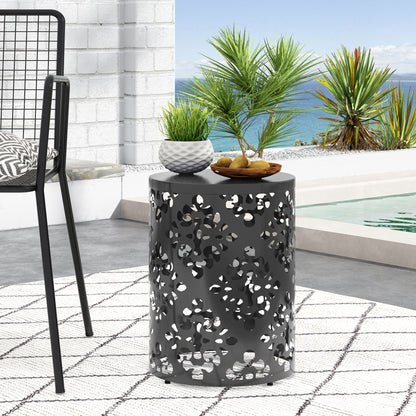 The Outdoor Metal Modern Design End Table, featuring a black finish and floral cutouts, sits on a white patterned outdoor rug beside a pool. Potted plants and fruit decorate the table, with ocean views and tropical plants in the background.