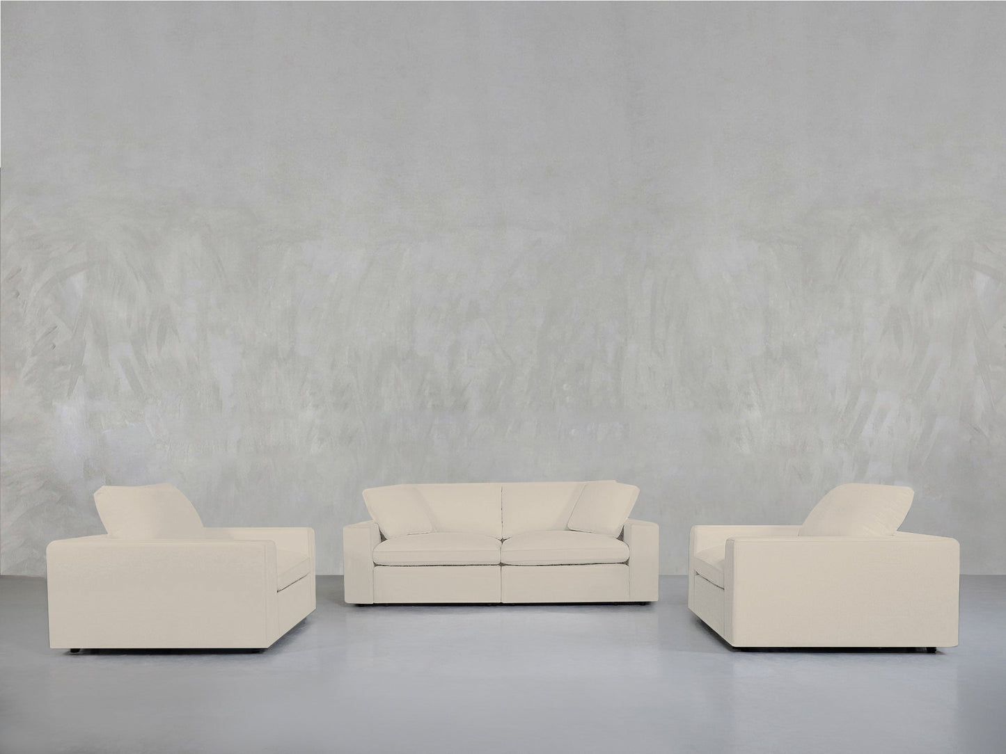 A minimalist room with a light grey textured wall features the 2-1-1 Sofa & Armchair Set in cream, arranged in a semi-circle on a smooth, light floor.