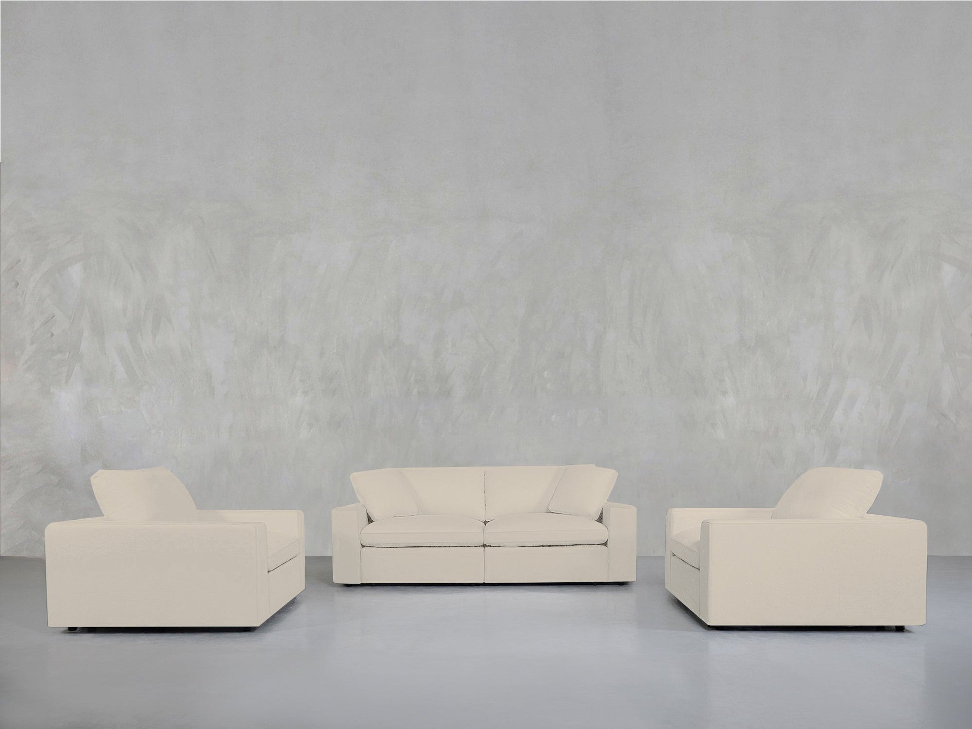 A minimalist room with a light grey textured wall features the 2-1-1 Sofa & Armchair Set in cream, arranged in a semi-circle on a smooth, light floor.