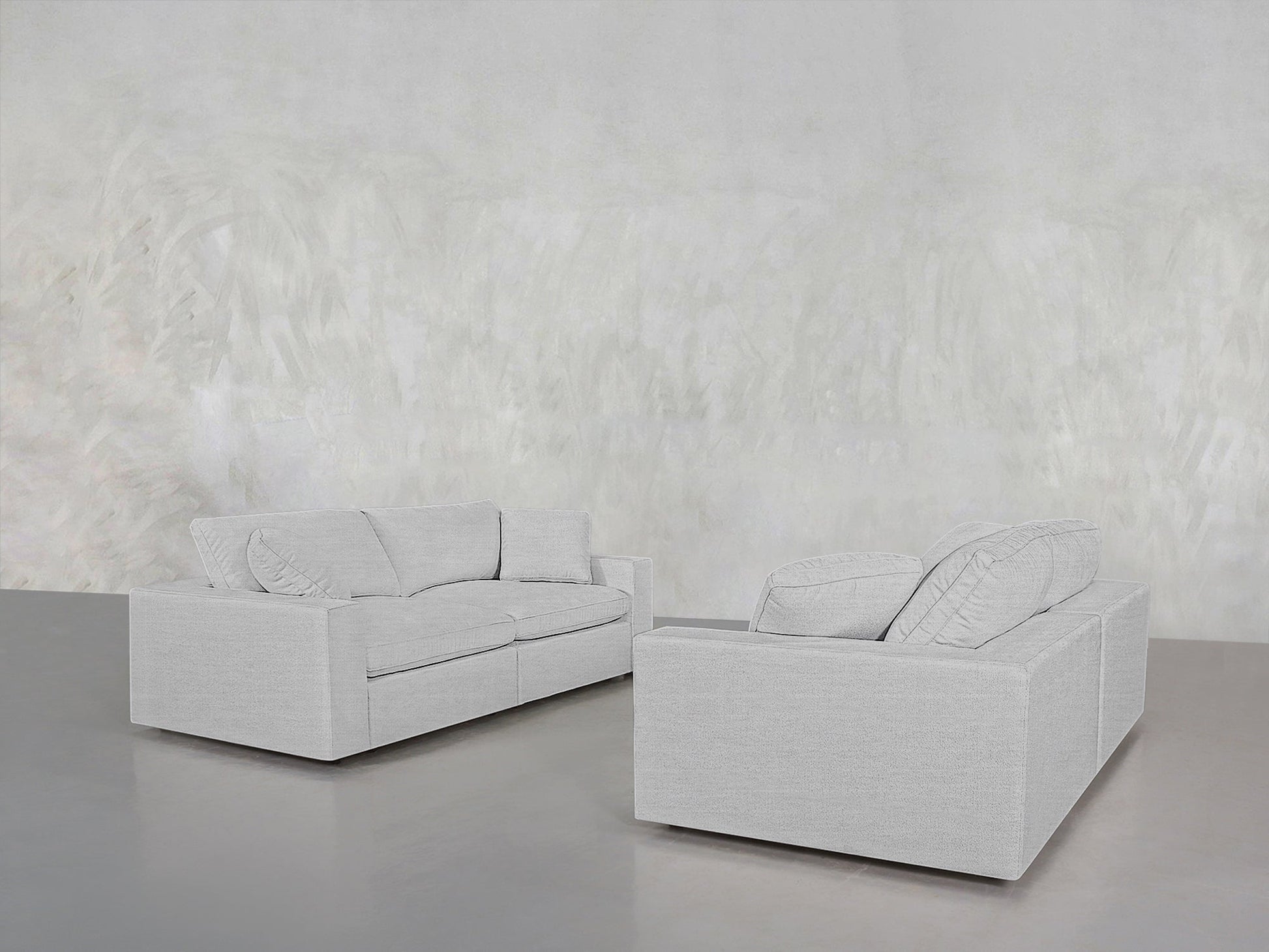 The 2-2 Sofa Set, featuring two light gray sofas with loose back cushions, is arranged facing each other in a minimalist room with a gray floor and a textured, light-colored wall.