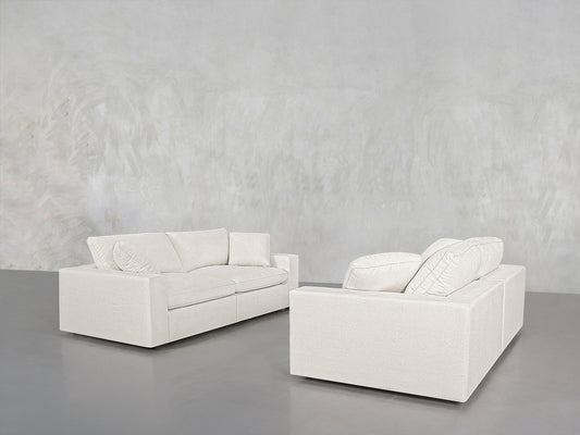 The 2-2 Sofa Set, featuring two modern white sofas with plush cushions, is arranged in a minimalistic room with a light grey textured wall and a smooth grey floor.
