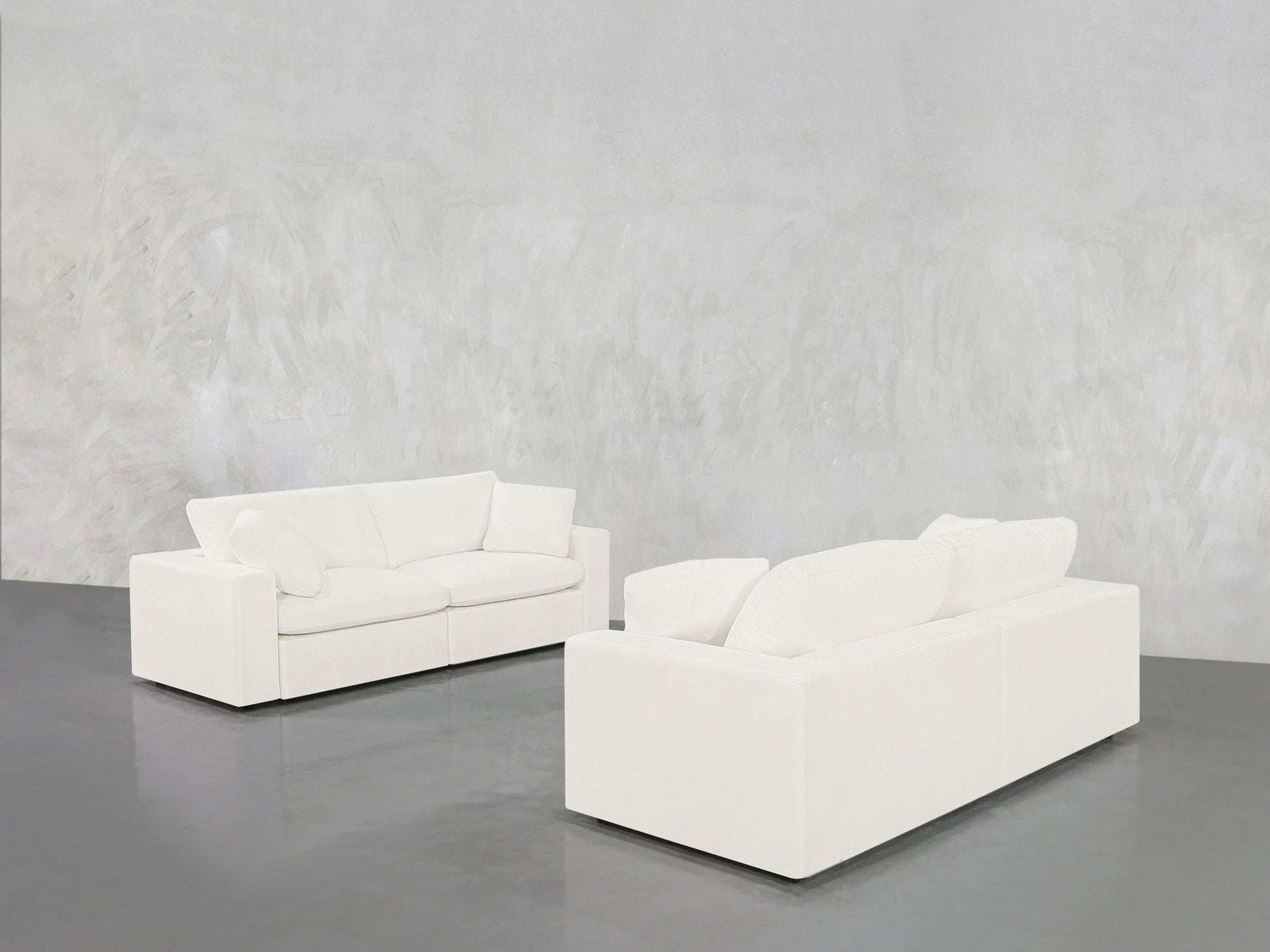 The 2-2 Sofa Set, with its modern white design and plush cushions, is arranged apart on a shiny gray floor against a textured light gray wall in a spacious, minimalist room.