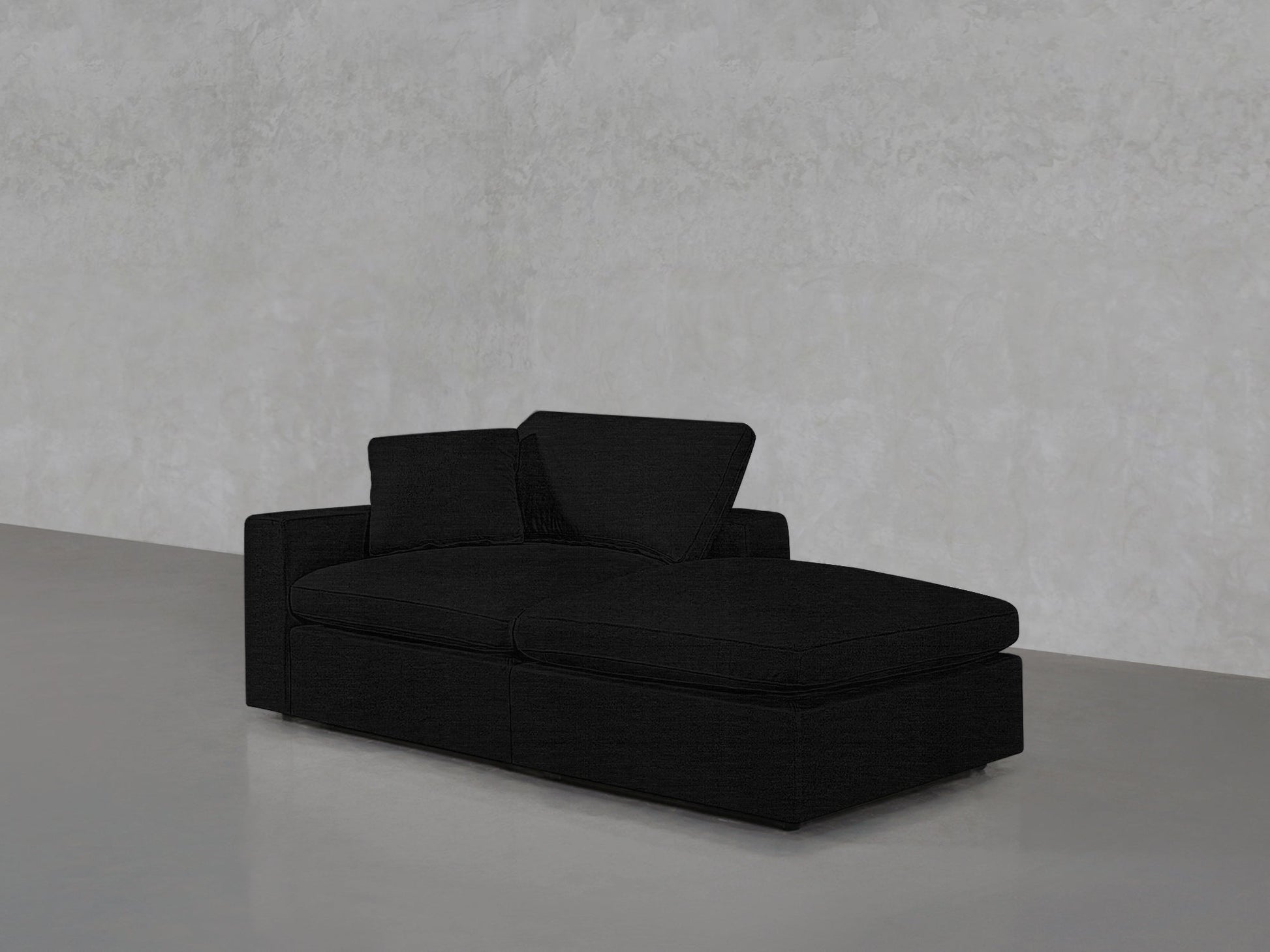 A 2-Seat Modular Lounger Loveseat in black, featuring two cushions and a chaise lounge, is set against a light gray textured wall on a smooth gray floor.
