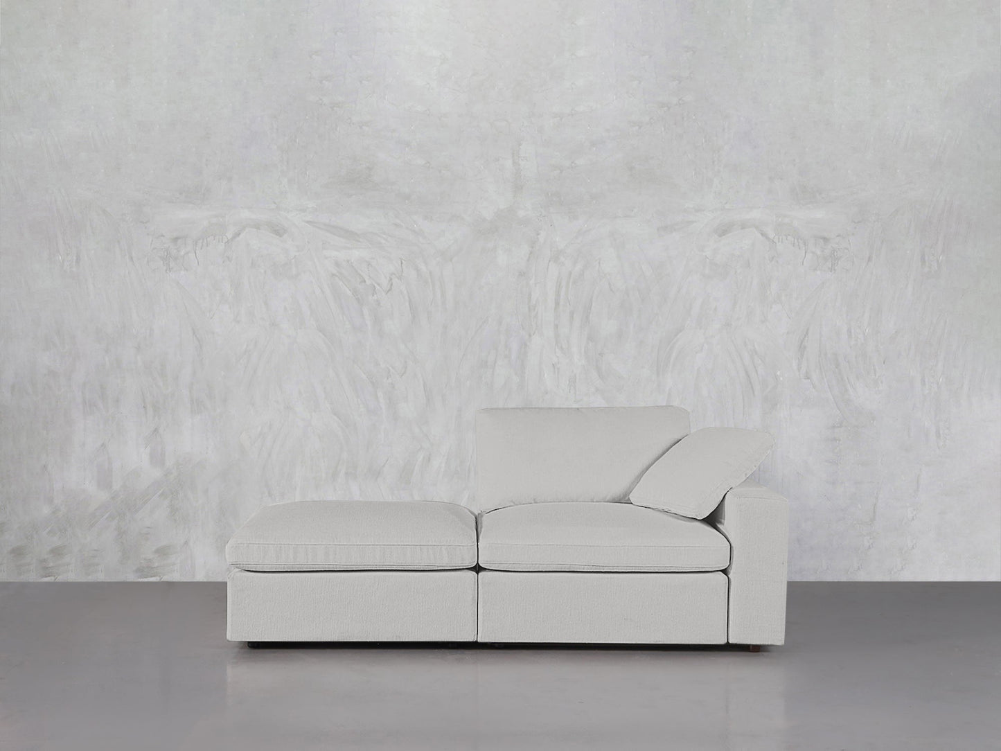 The 2-Seat Modular Lounger Loveseat, in light gray with one armrest and a chaise lounge, is placed on a smooth gray floor by a textured white wall.