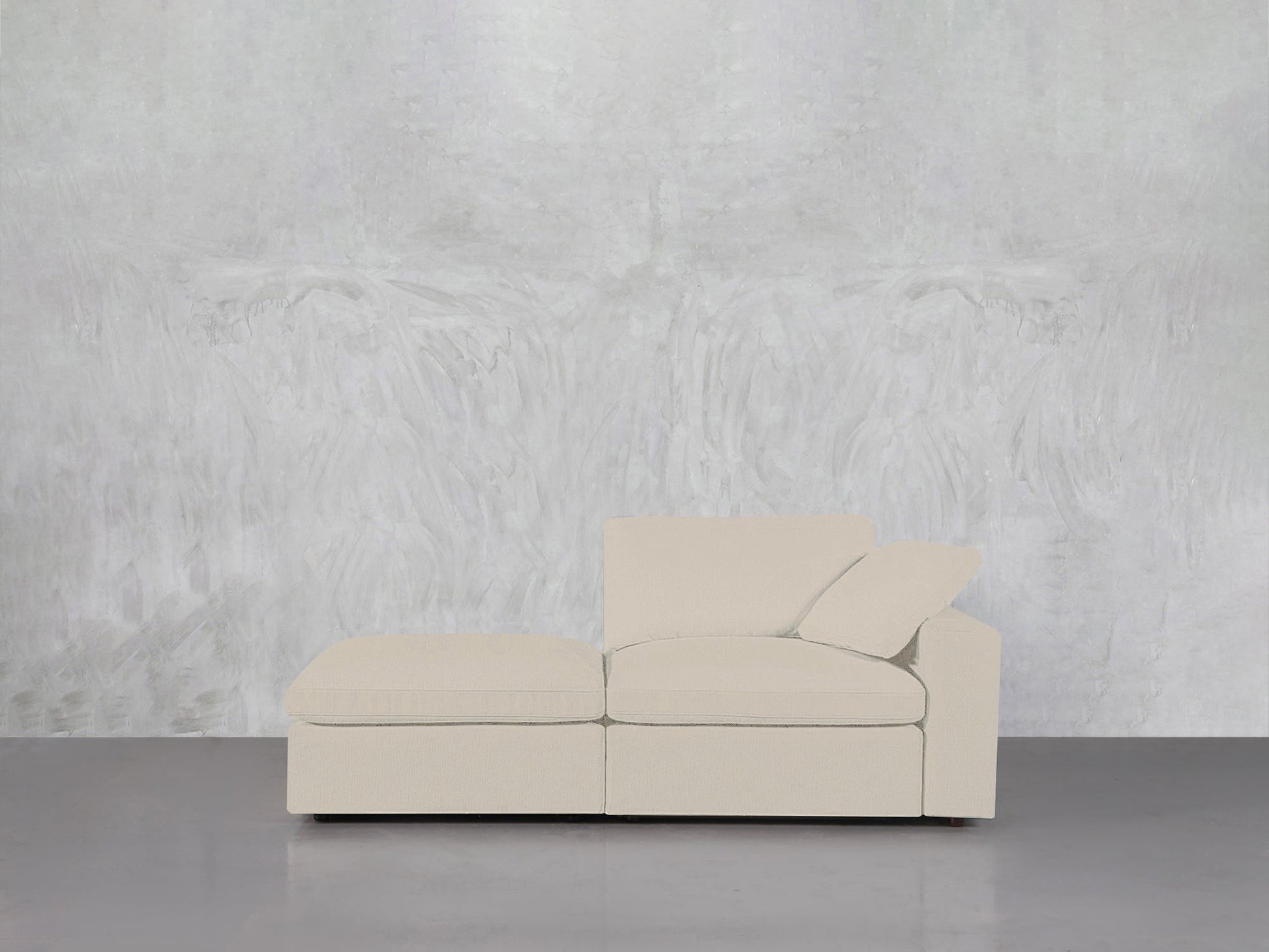 A contemporary 2-Seat Modular Lounger Loveseat in light beige with a chaise lounge sits on a gray floor against a textured light gray wall, featuring one matching cushion on the right side.