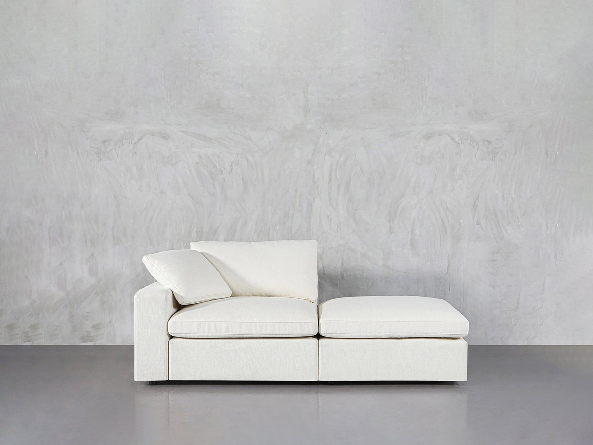 The 2-Seat Modular Lounger Loveseat, featuring clean lines and plush off-white cushions, sits against a textured light grey wall on a smooth grey floor.