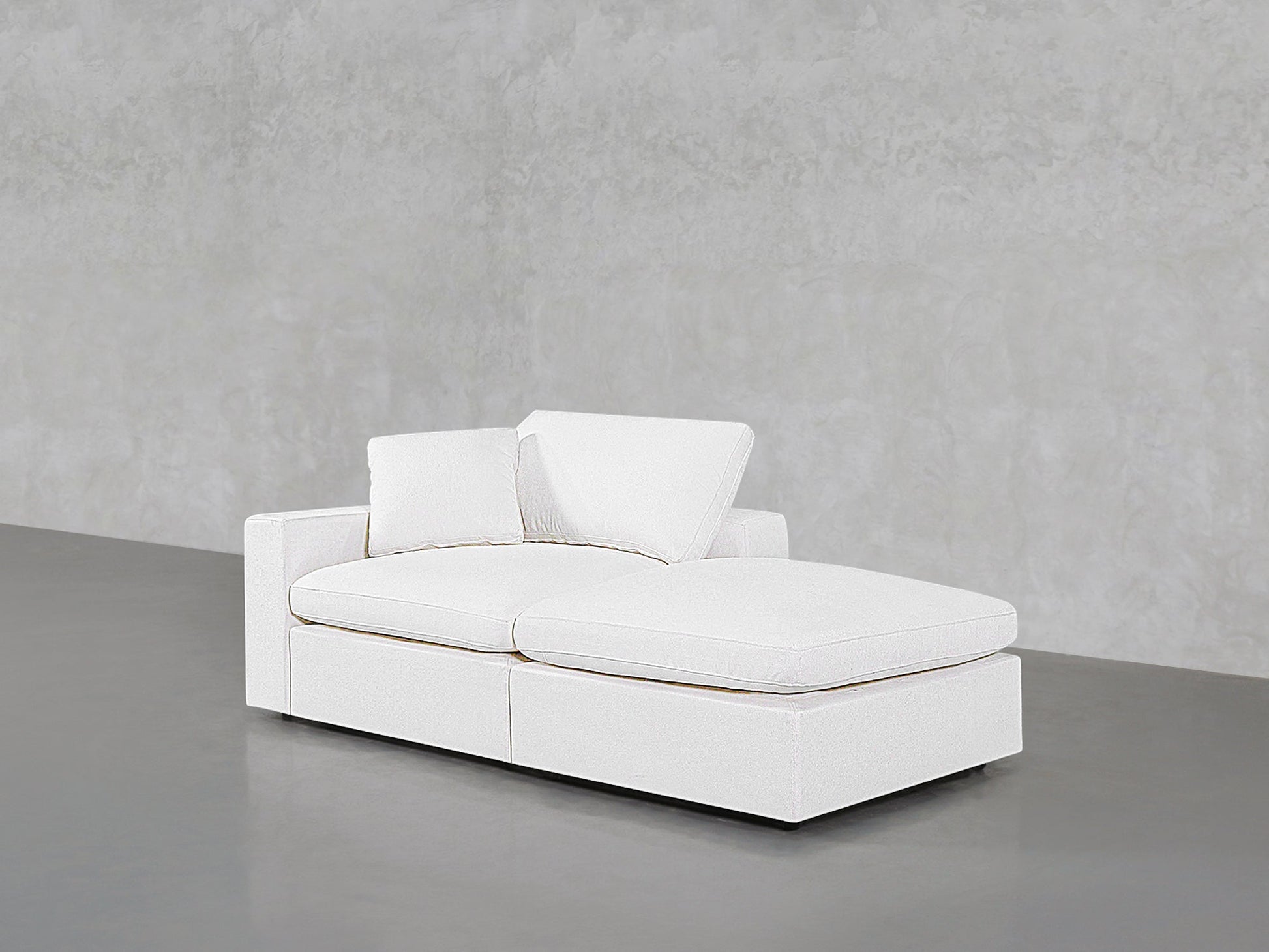 A 2-Seat Modular Lounger Loveseat in modern white upholstery with two matching cushions is placed on a smooth grey floor against a textured light grey wall.