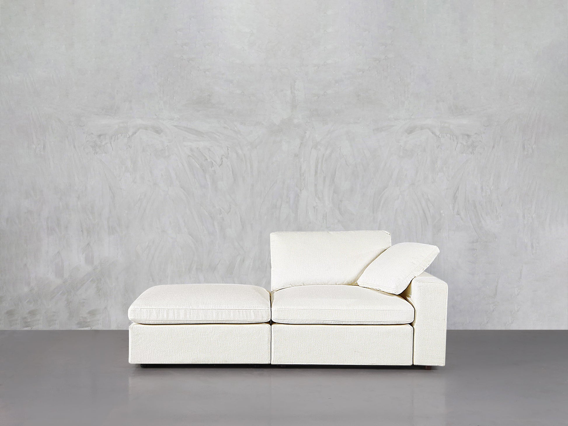 The 2-Seat Modular Lounger Loveseat in off-white, featuring a right armrest and loose cushion, sits against a light gray textured wall on a smooth gray floor.