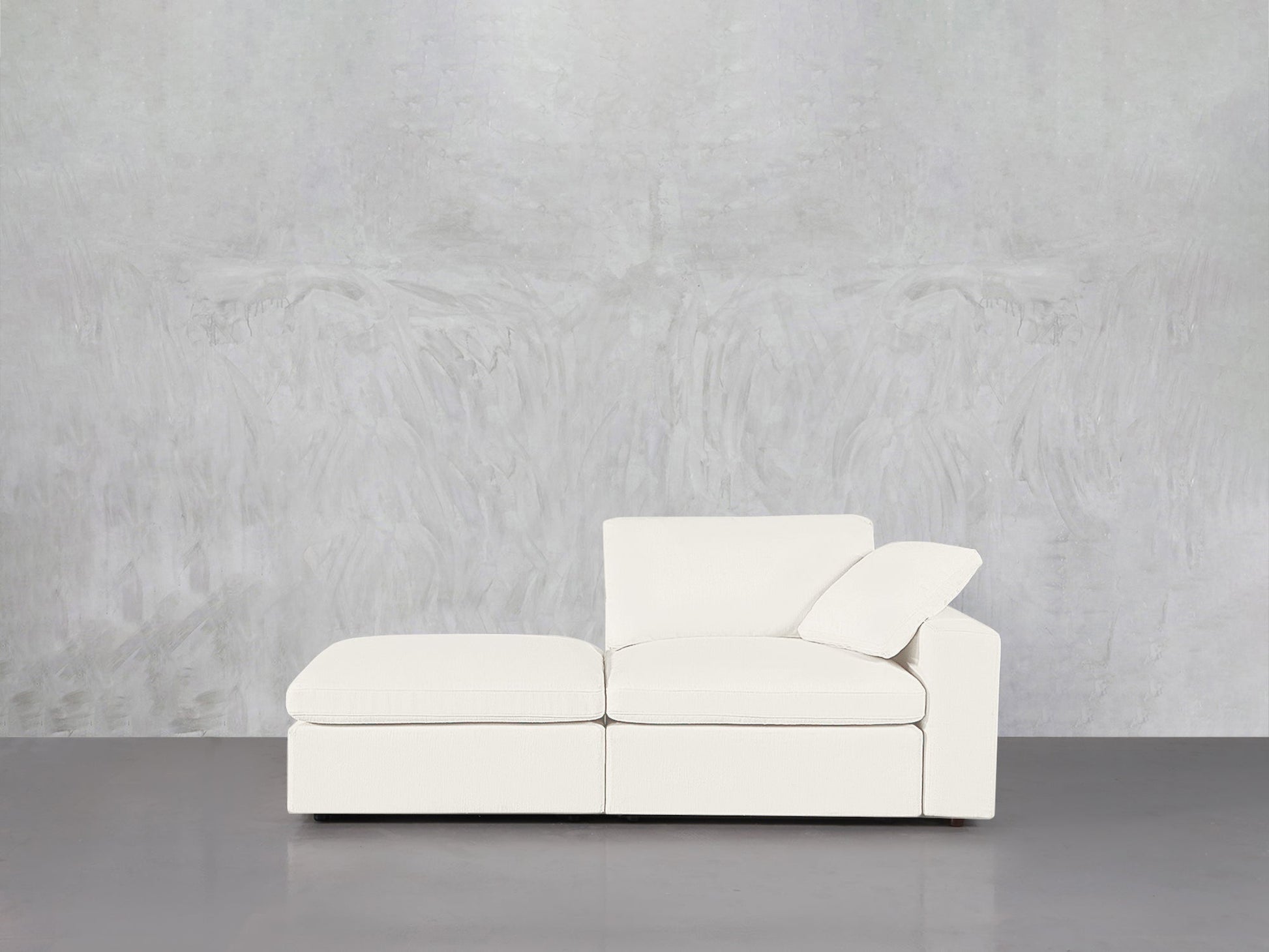 A 2-Seat Modular Lounger Loveseat in white, paired with an ottoman, sits against a light gray textured wall on a smooth gray floor. One cushion is angled for support in this minimalistic, uncluttered space.