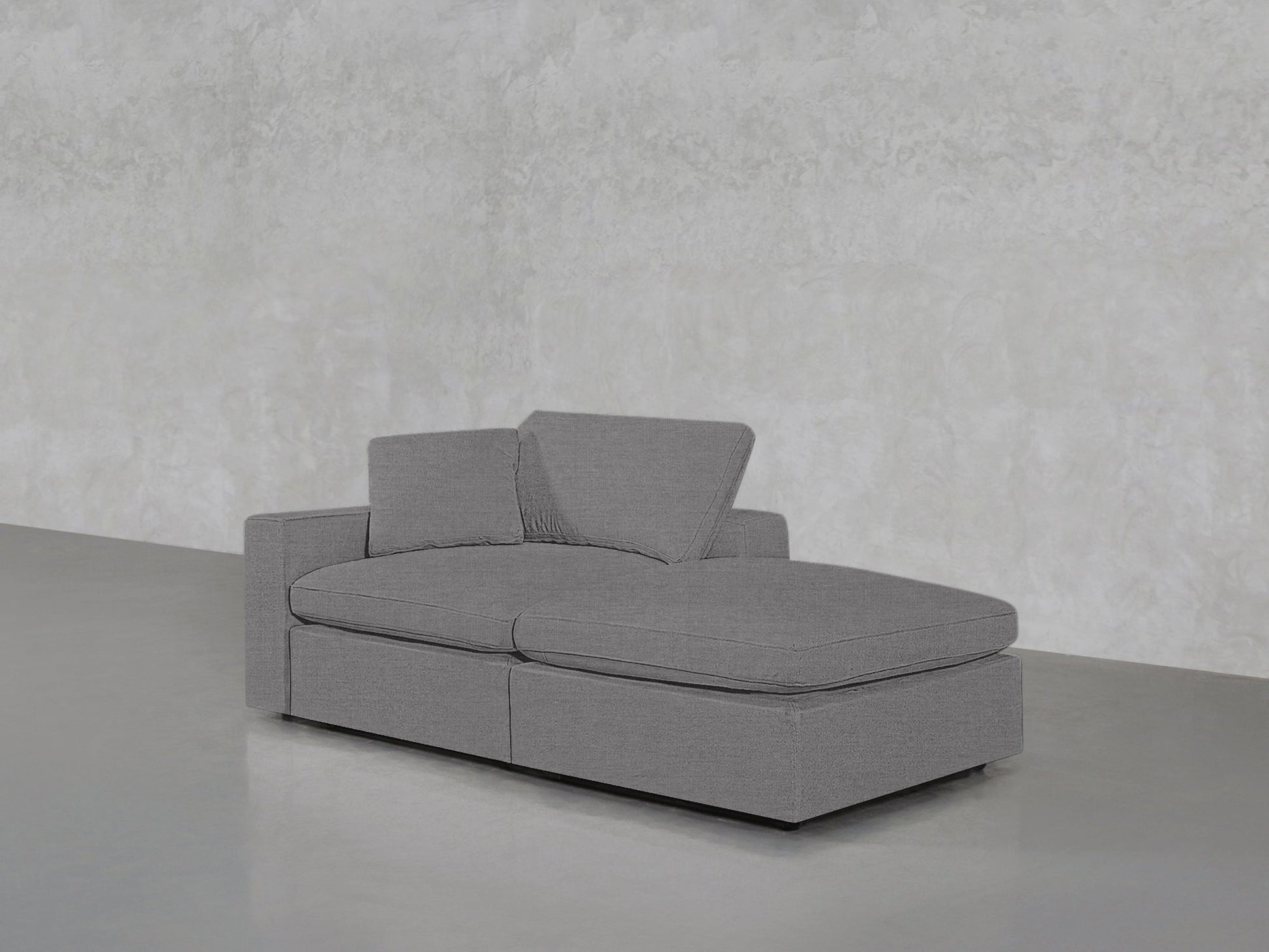 The 2-Seat Modular Lounger Loveseat, in modern gray with two seat and back cushions plus an extended chaise, sits on a smooth gray floor against a textured light gray wall.