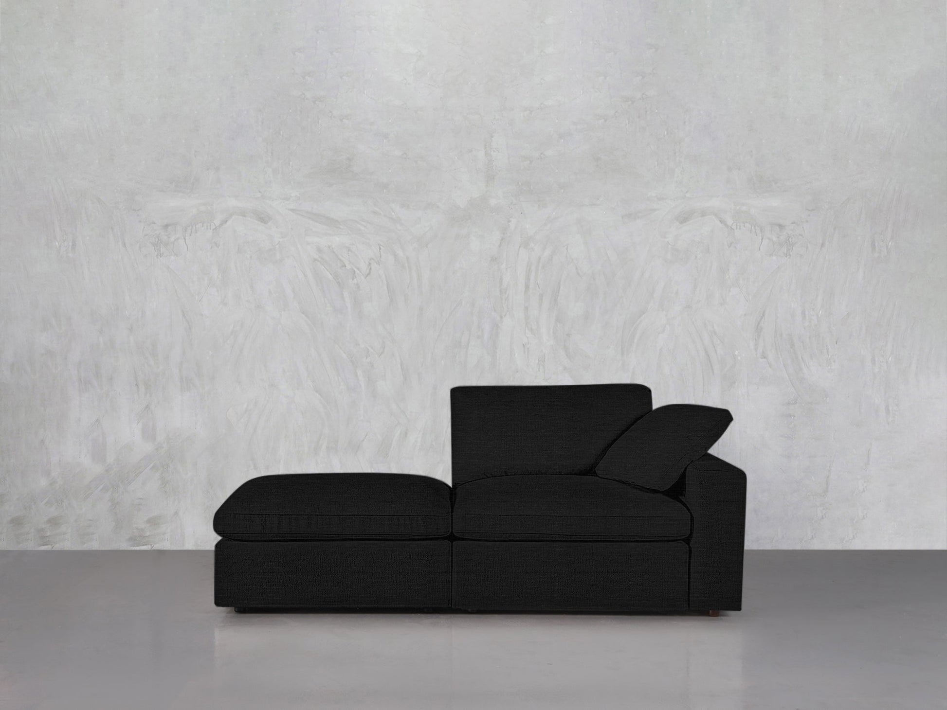 A black 2-Seat Modular Lounger Loveseat with a chaise sits against a light gray textured wall on a glossy gray floor. The minimalist room features only the loveseat, emphasizing its modern design.