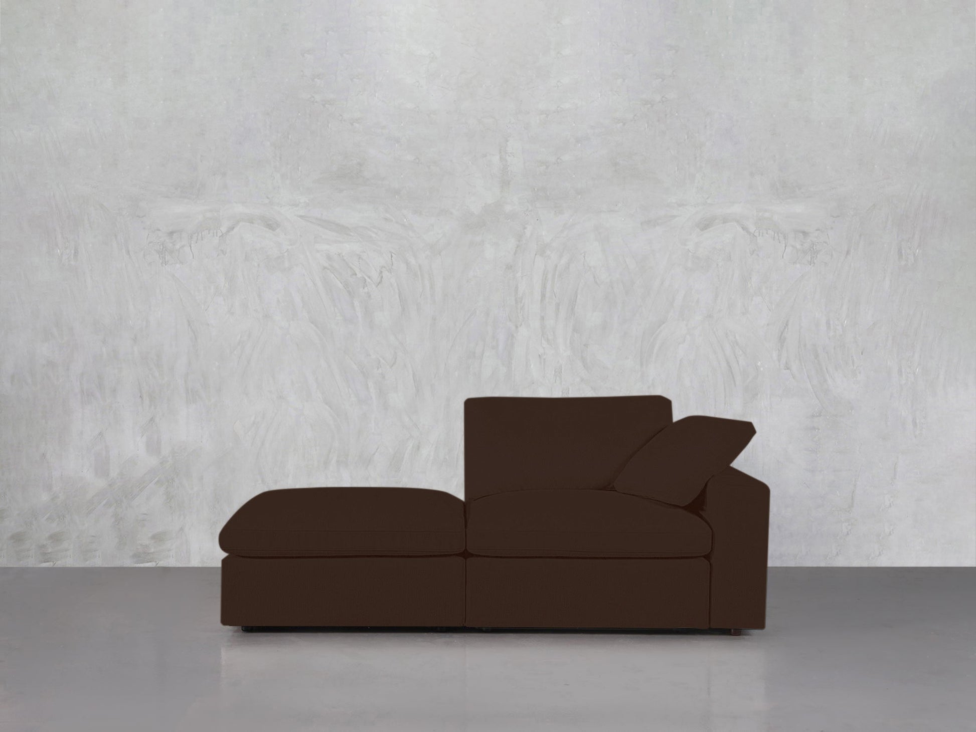 The 2-Seat Modular Lounger Loveseat in dark brown stands against a textured light gray wall on a smooth gray floor. One cushion is angled for back support, creating a minimalist and uncluttered setting.