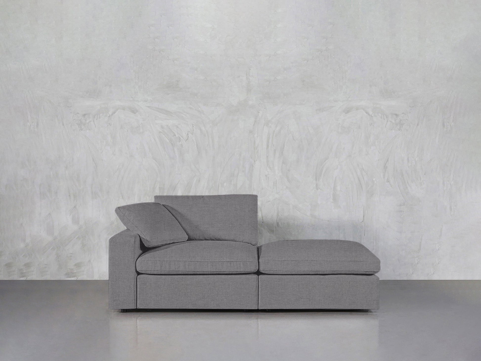 A 2-Seat Modular Lounger Loveseat in modern gray with two seat cushions and back pillows is placed against a textured light gray wall on a smooth gray floor.