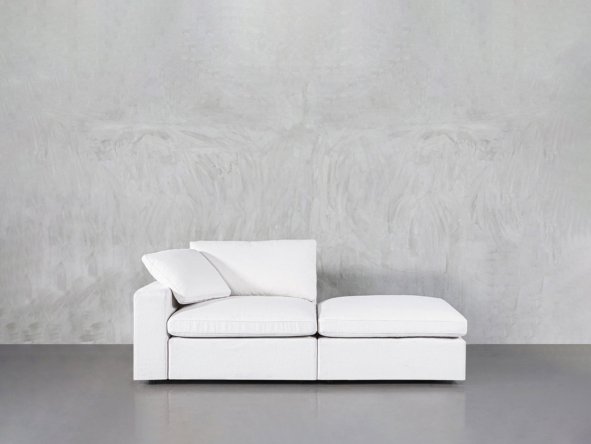A 2-Seat Modular Lounger Loveseat with two cushions is placed against a light gray textured wall on a smooth gray floor.