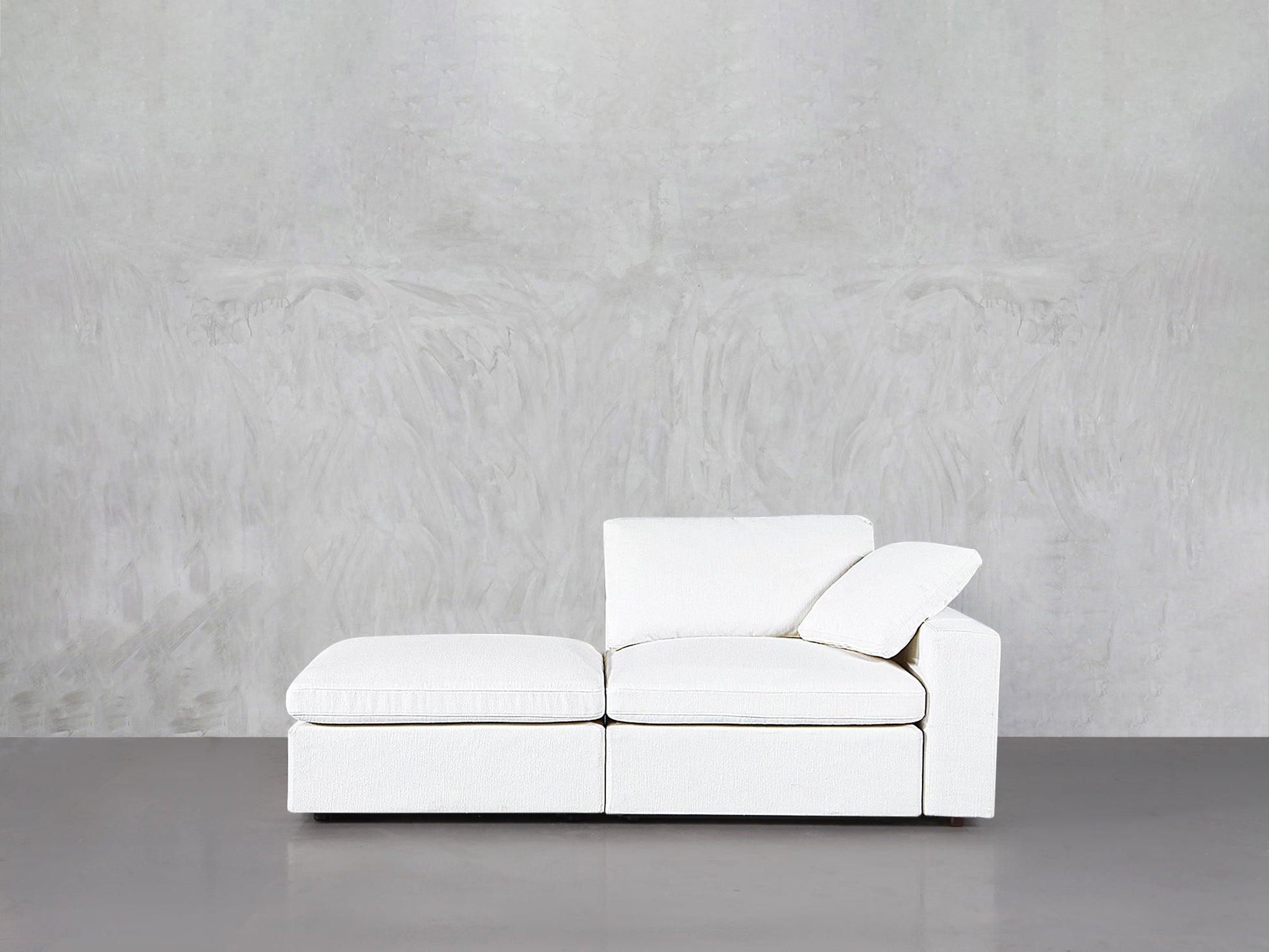 A 2-Seat Modular Lounger Loveseat in modern white with clean lines sits against a light gray textured wall on a smooth gray floor, featuring one armrest and a single back cushion.
