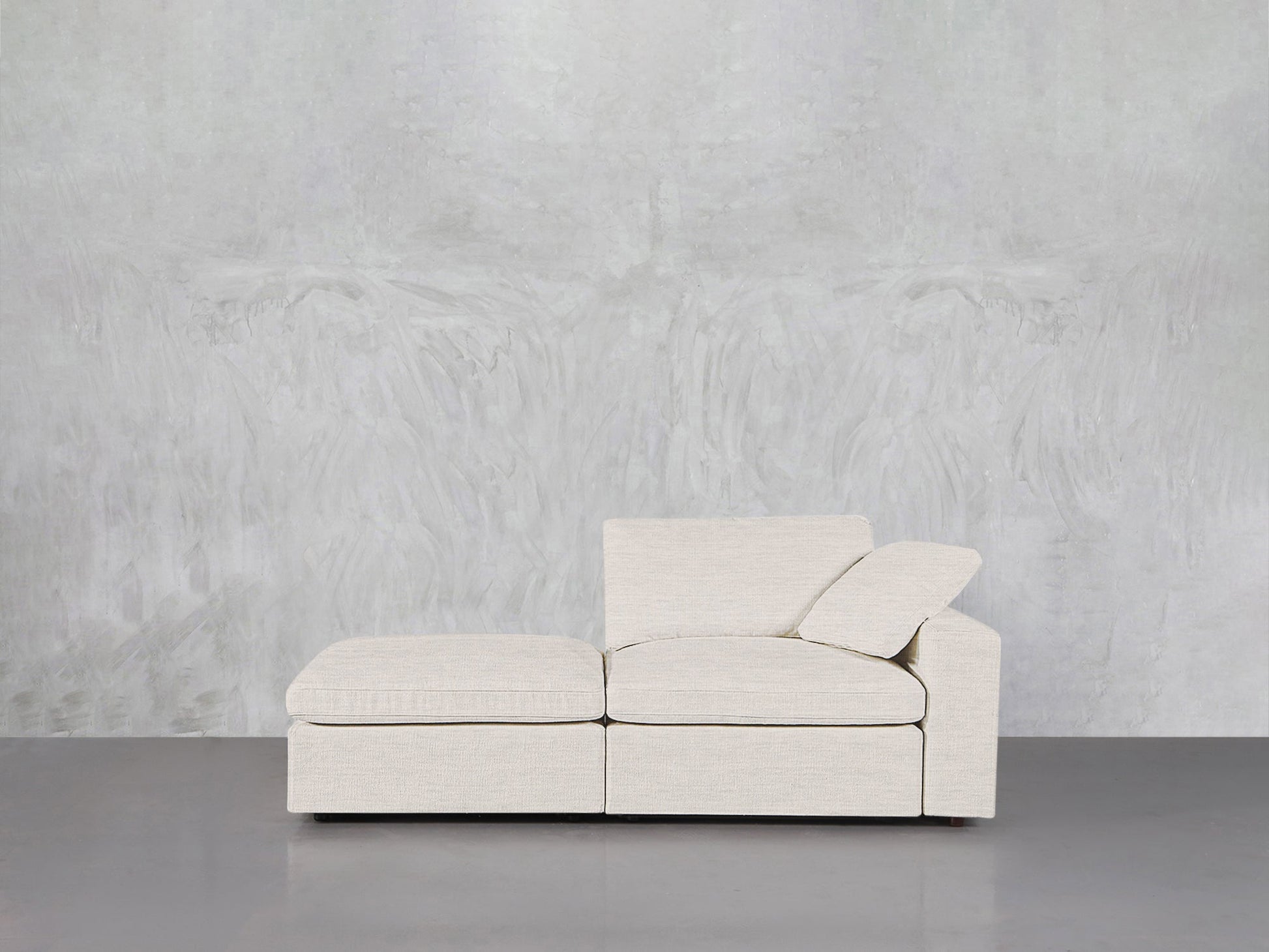 The 2-Seat Modular Lounger Loveseat in light beige, featuring an ottoman and angled backrest, sits before a textured light gray wall on a smooth gray floor.